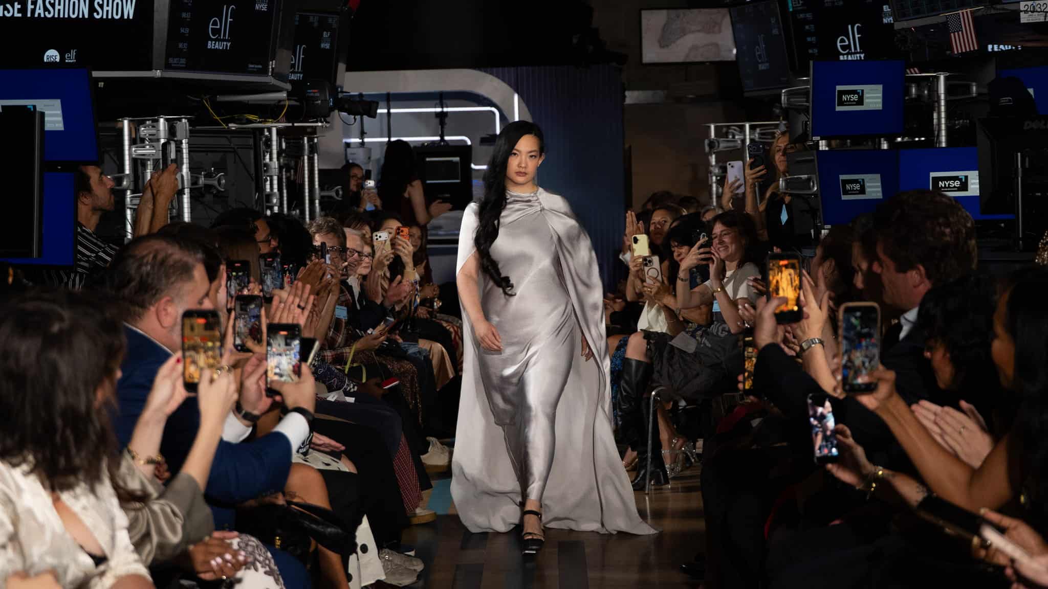 A model in a flowing silver dress walks down a runway, surrounded by a crowd of seated spectators, many holding up phones to take photos and videos during a fashion show.