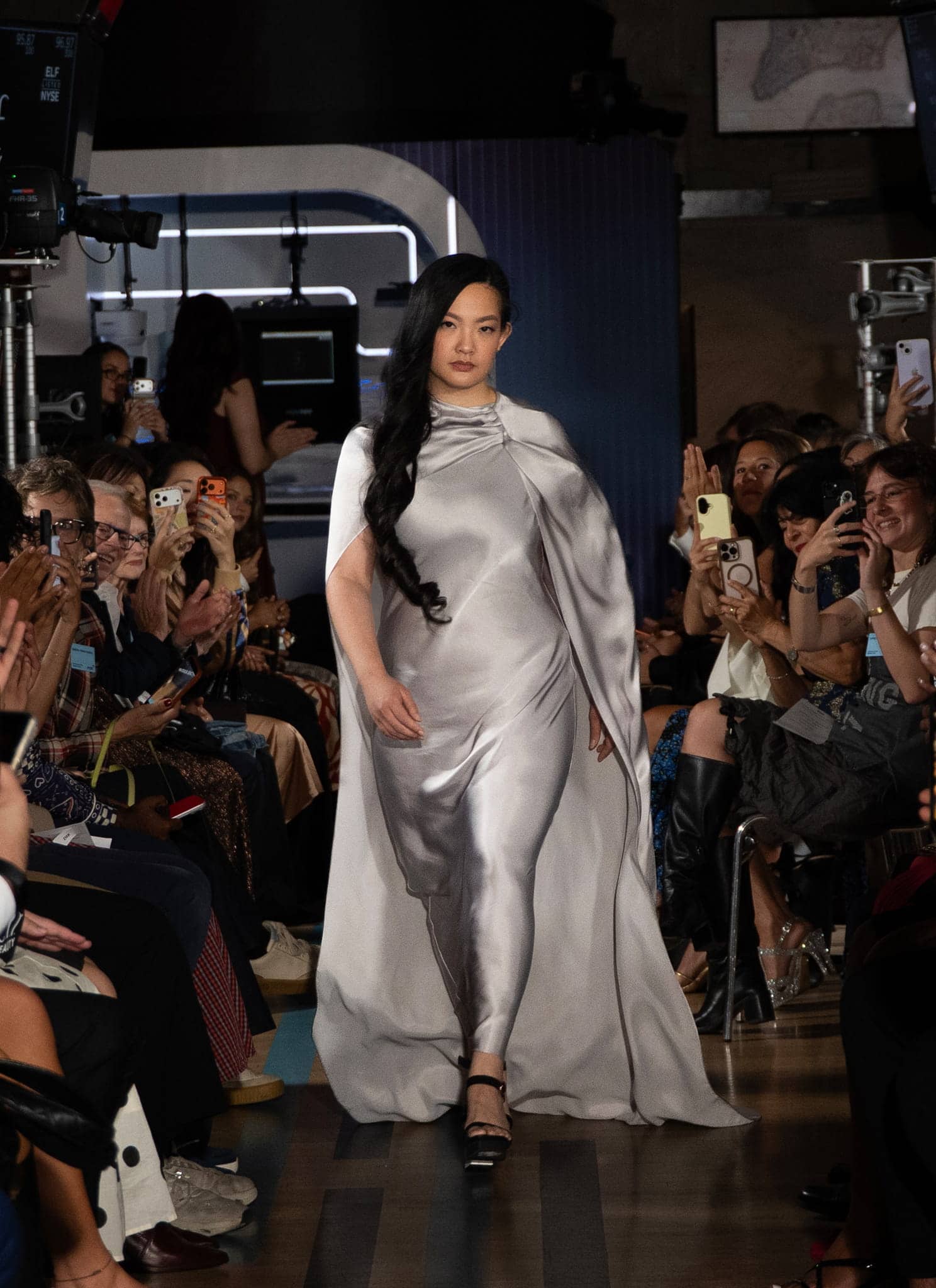 A model in a flowing silver dress walks down a runway, surrounded by a crowd of seated spectators, many holding up phones to take photos and videos during a fashion show.