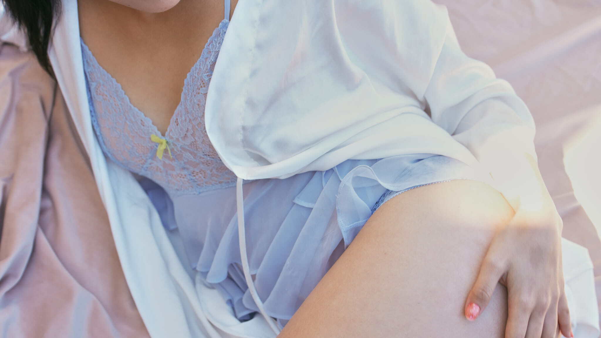 A woman lying on light-colored bedding, wearing a blue lace-trimmed slip and a white robe. One hand rests on her thigh, and the lighting is soft and warm.