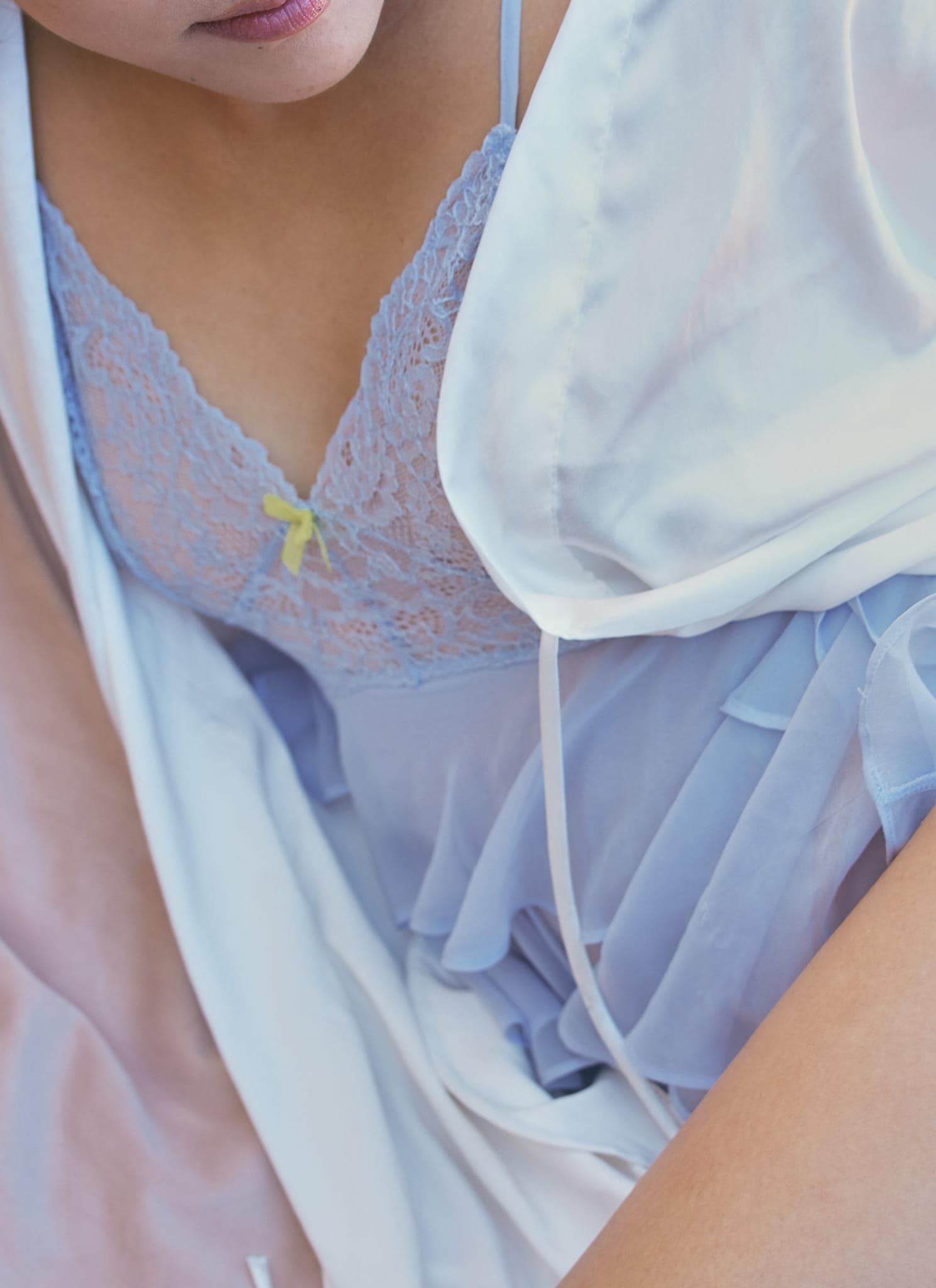 A woman lying on light-colored bedding, wearing a blue lace-trimmed slip and a white robe. One hand rests on her thigh, and the lighting is soft and warm.