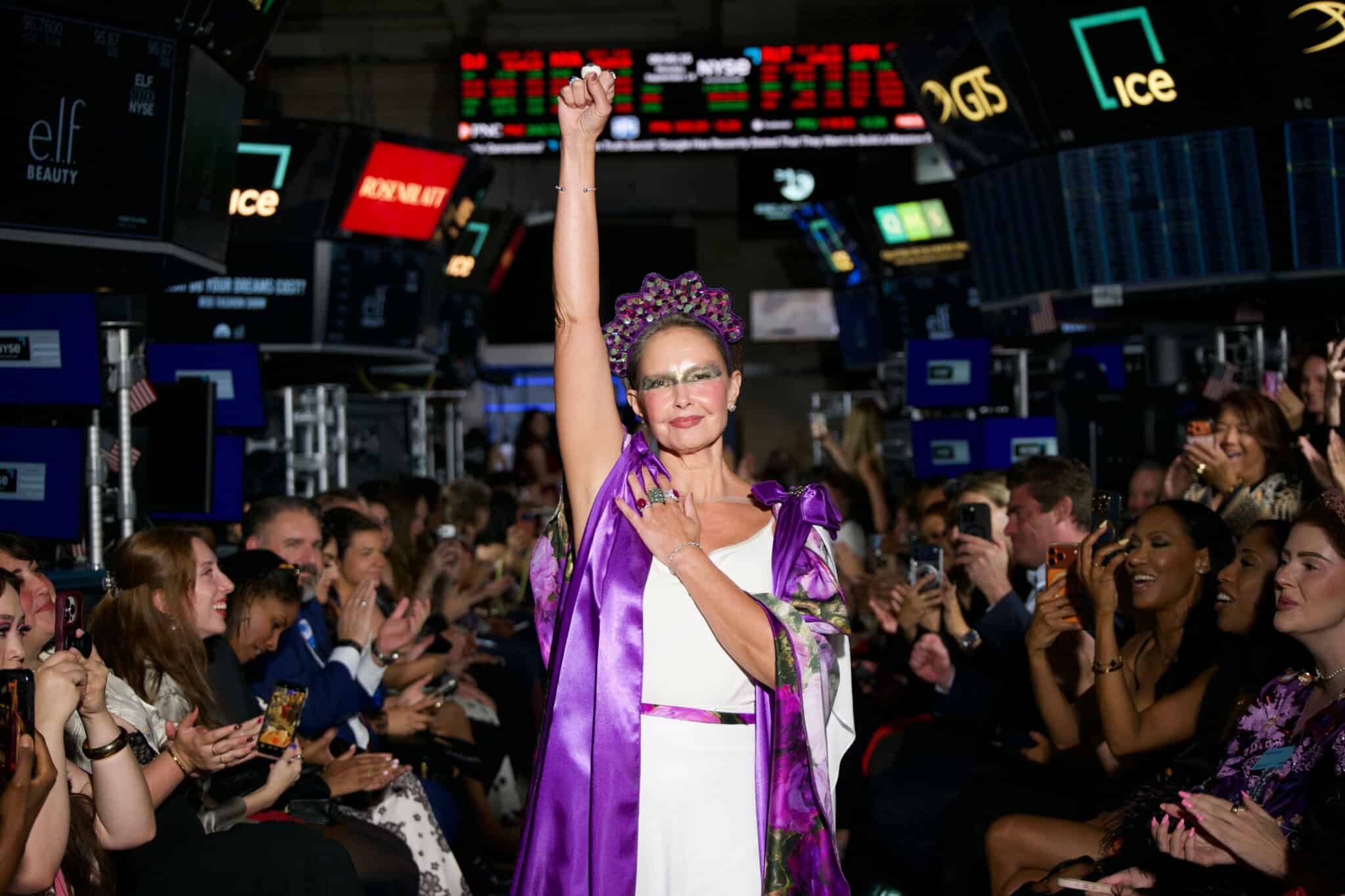 A woman in a crown and purple cape raises her fist on a runway, surrounded by cheering people. Stock market tickers and monitors are visible in the background.