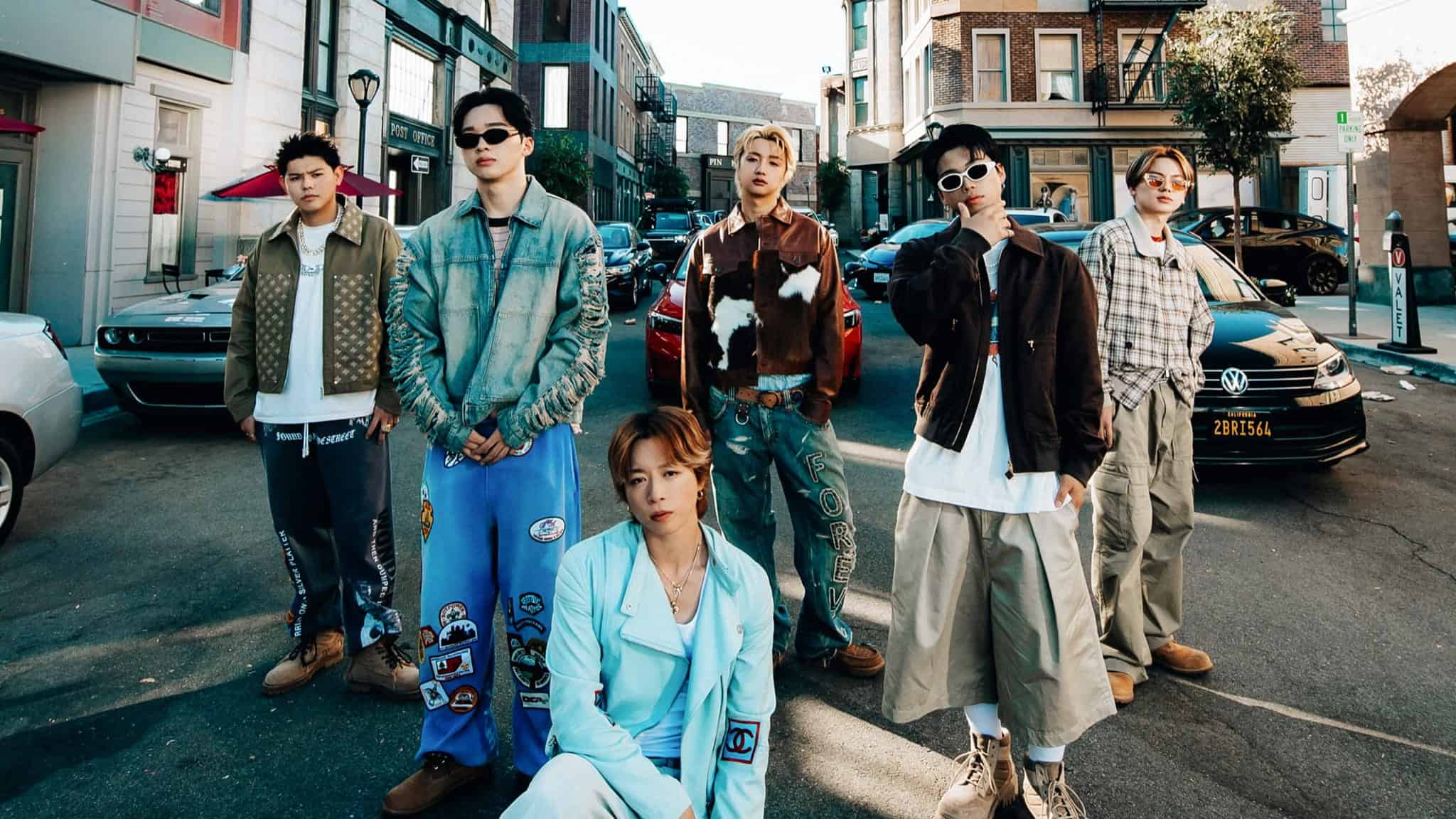 Six young men in stylish, casual streetwear pose confidently in the middle of a city street, with cars and buildings in the background.
