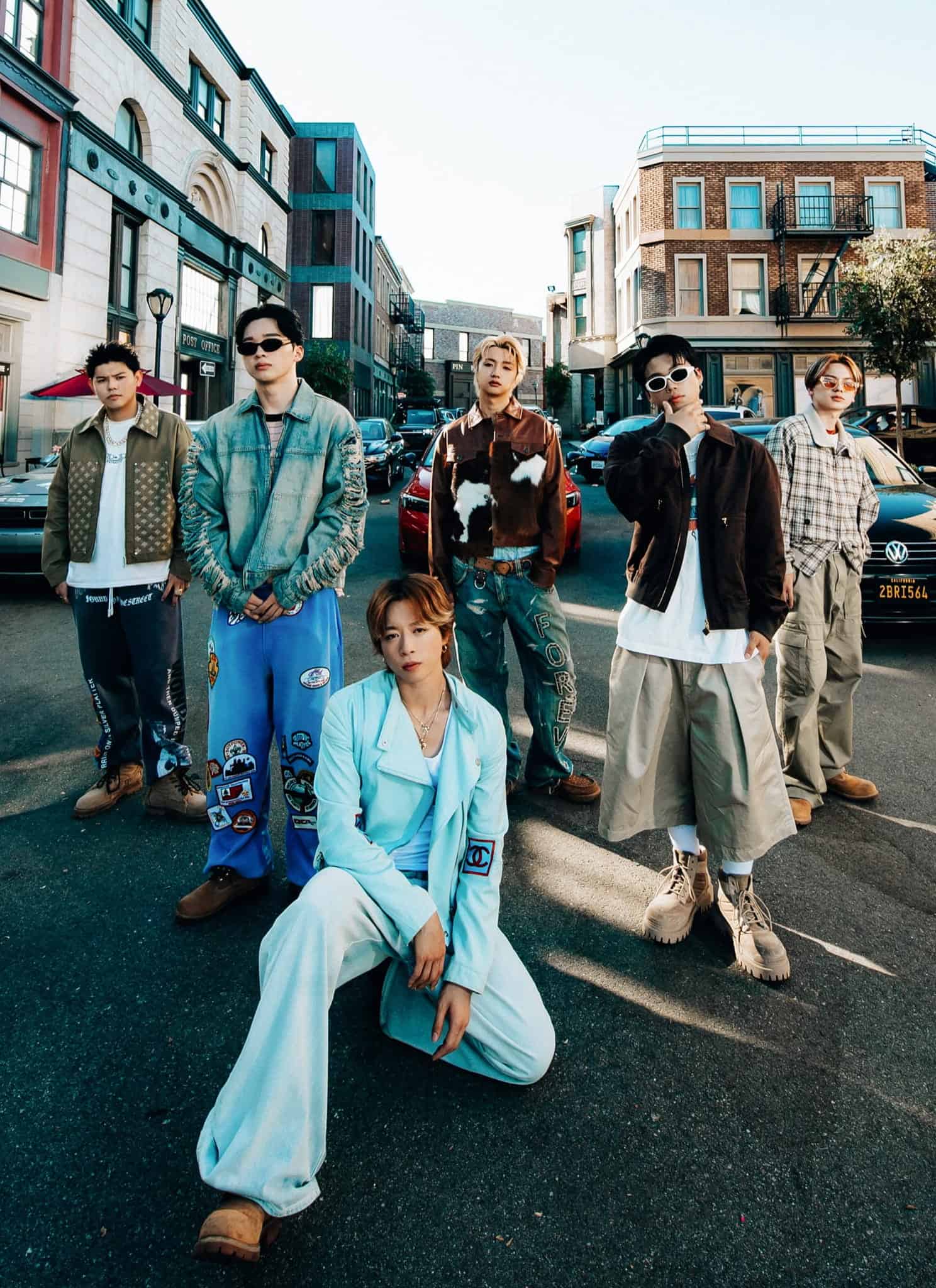 Six young men in stylish, casual streetwear pose confidently in the middle of a city street, with cars and buildings in the background.