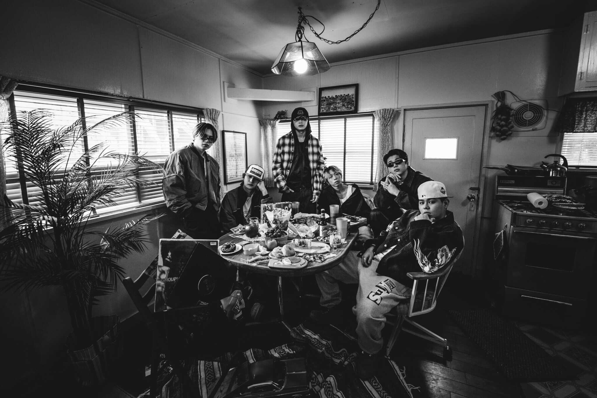 A black-and-white photo of six people posing around a cluttered kitchen table with food and drinks. The setting has a casual, relaxed atmosphere, with some people standing and others sitting.