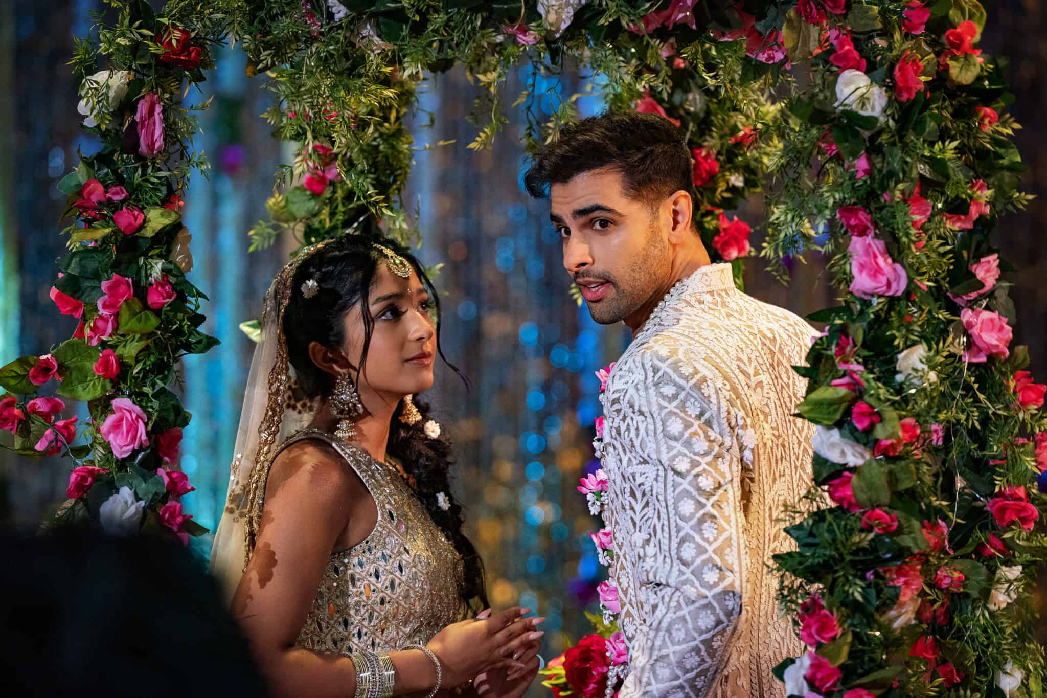 A couple in ornate traditional attire stands under a floral arch, looking at each other. The setting is festive, with vibrant pink and green flowers and shimmering decorations in the background.