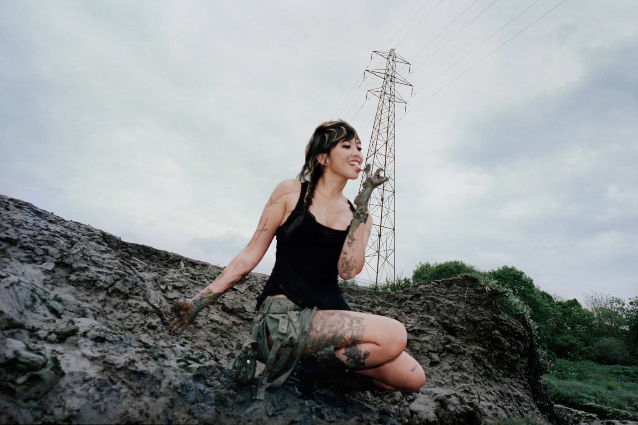 A woman with braids, wearing a black tank top and muddy shorts, kneels on a muddy hill, smiling with mud on her hands and legs. Power lines and a cloudy sky are visible in the background.