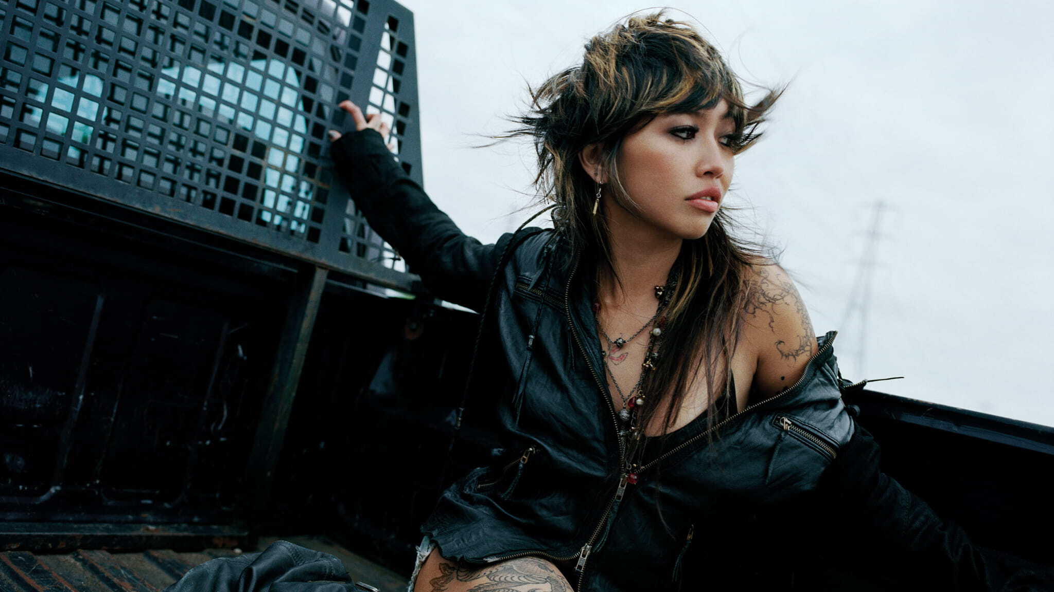 A woman with layered hair, tattoos, and a black leather jacket sits in the back of a truck, looking to the side with a thoughtful expression. The sky is overcast and theres a metal grate behind her.