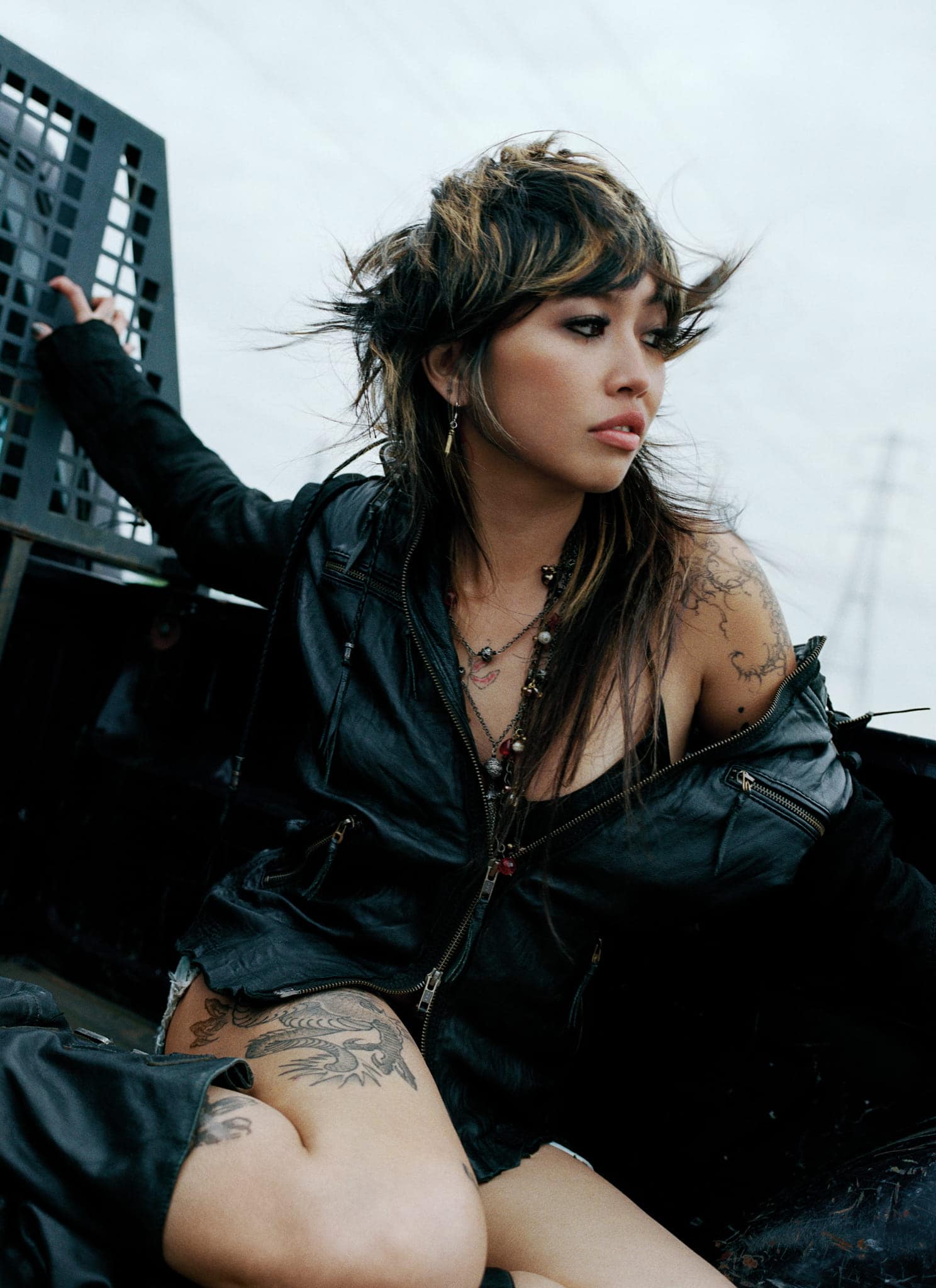 A woman with layered hair, tattoos, and a black leather jacket sits in the back of a truck, looking to the side with a thoughtful expression. The sky is overcast and theres a metal grate behind her.