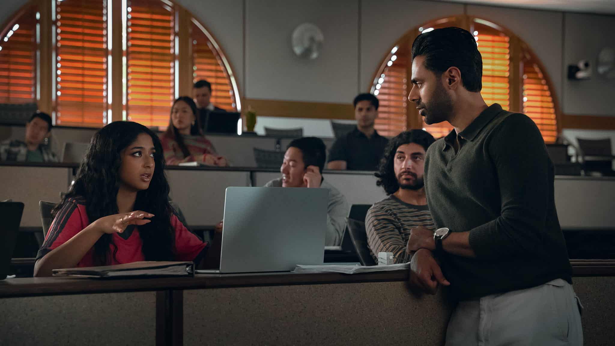 A woman gestures while speaking to a man standing beside her in a classroom. Other students sit in the background, some watching the conversation, with laptops and notebooks on desks.