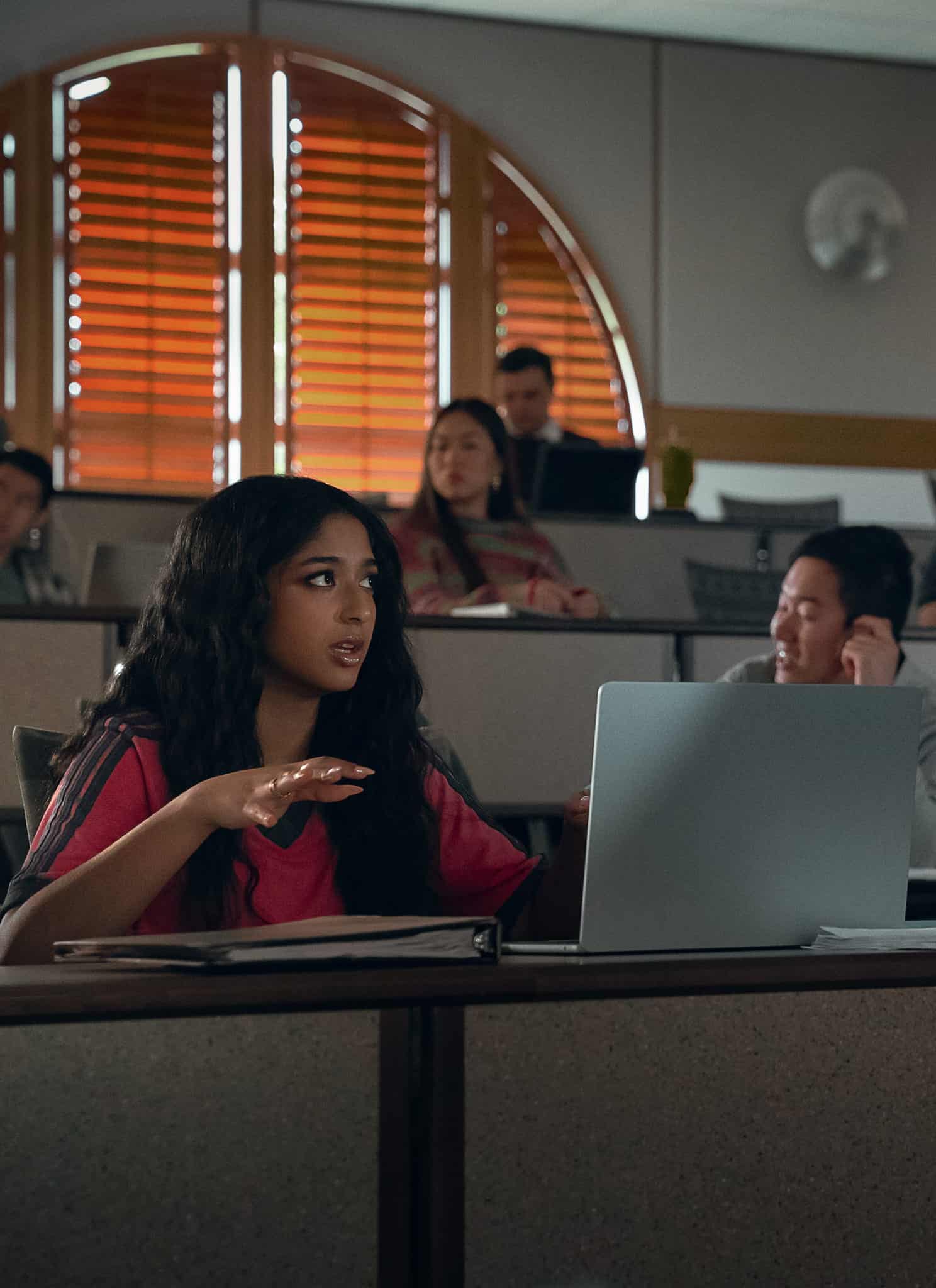 A woman gestures while speaking to a man standing beside her in a classroom. Other students sit in the background, some watching the conversation, with laptops and notebooks on desks.