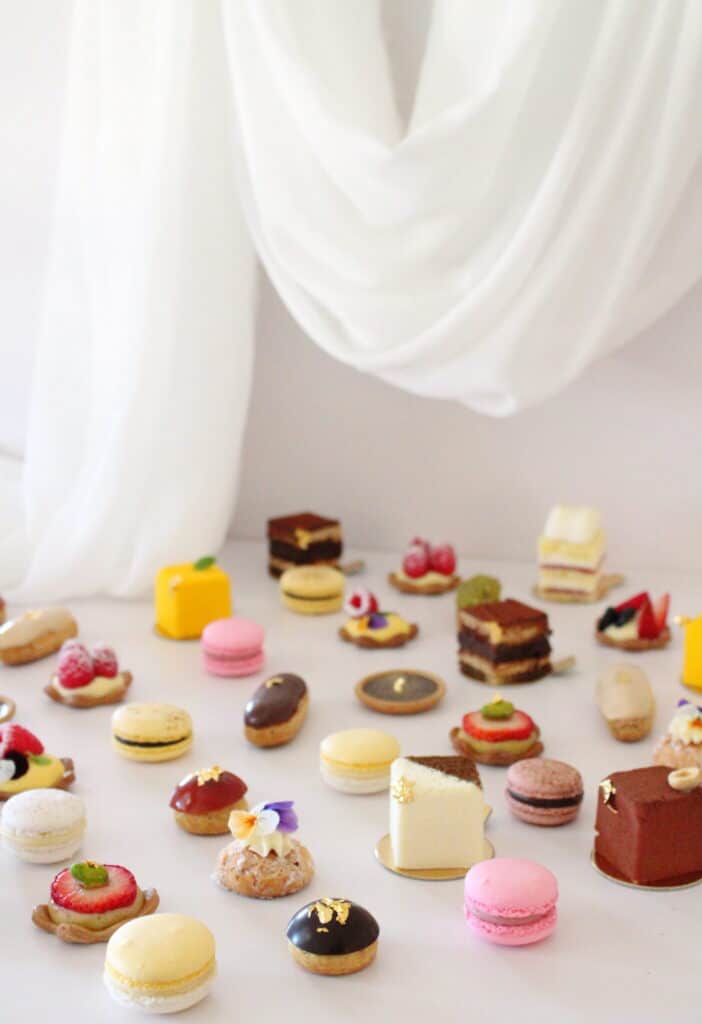 An assortment of colorful pastries and desserts, including macarons, cakes, and tarts, is arranged on a white surface with soft white fabric draped in the background.
