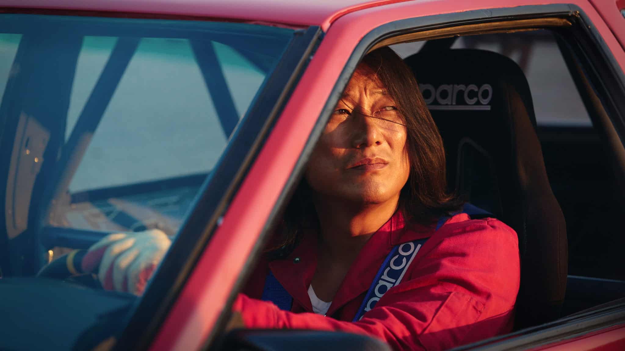 A person wearing a red jacket and racing harness sits in a red car, looking thoughtfully out the window. Sunlight illuminates their face, and the Sparco racing seat is visible.