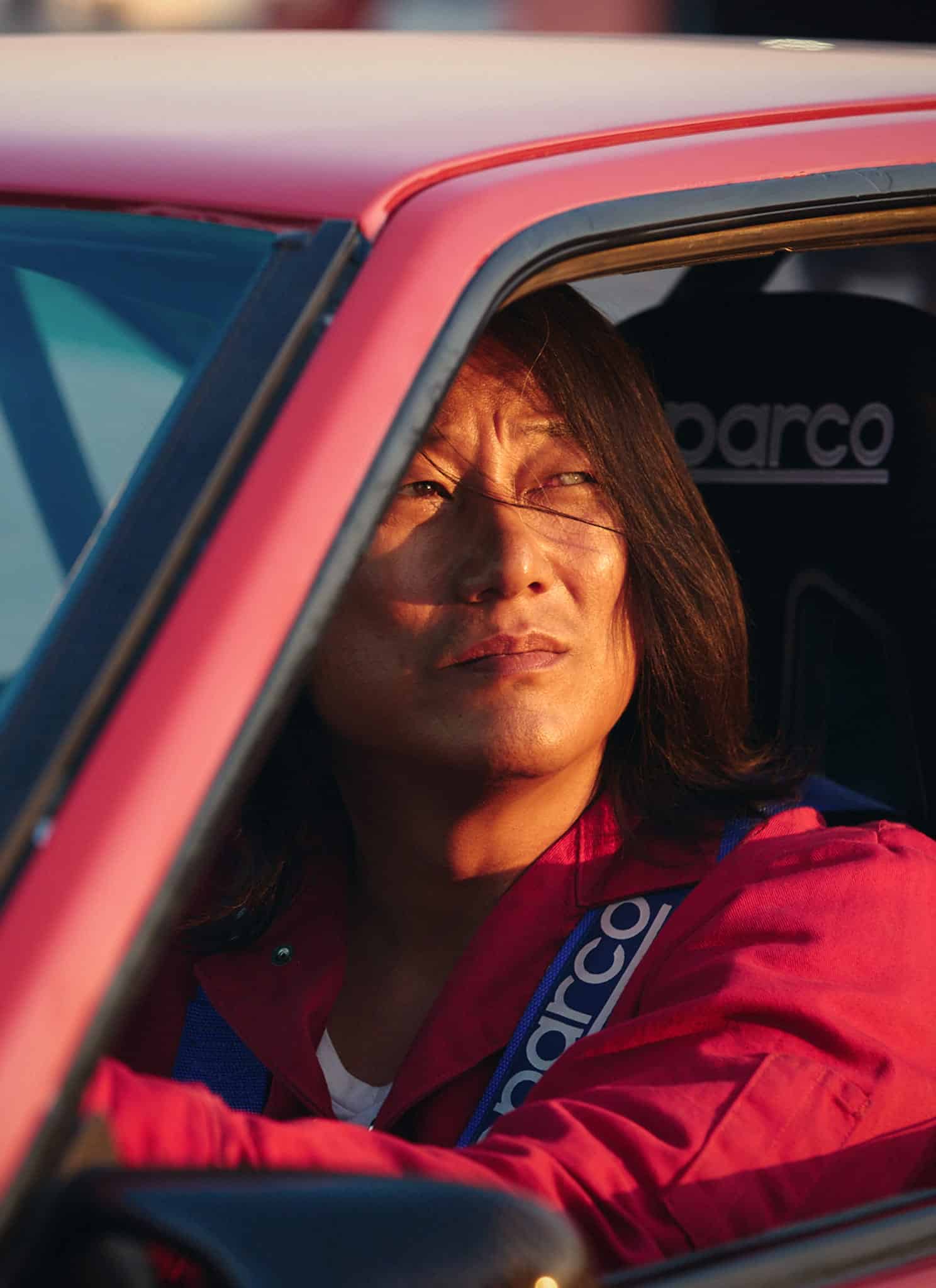 A person wearing a red jacket and racing harness sits in a red car, looking thoughtfully out the window. Sunlight illuminates their face, and the Sparco racing seat is visible.