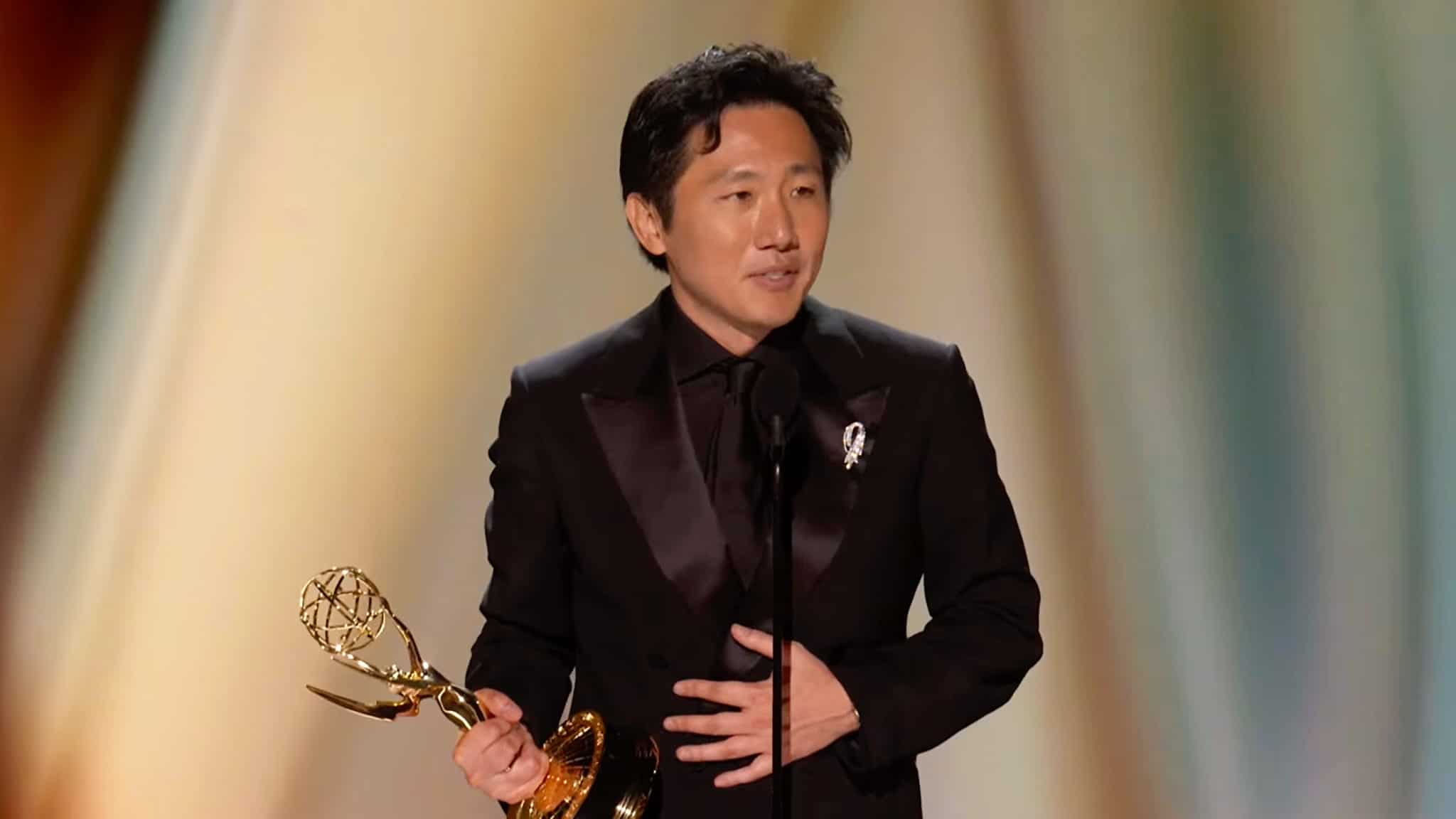 A man in a black suit stands on stage holding an Emmy award, speaking into a microphone against a softly blurred, gold-toned background.
