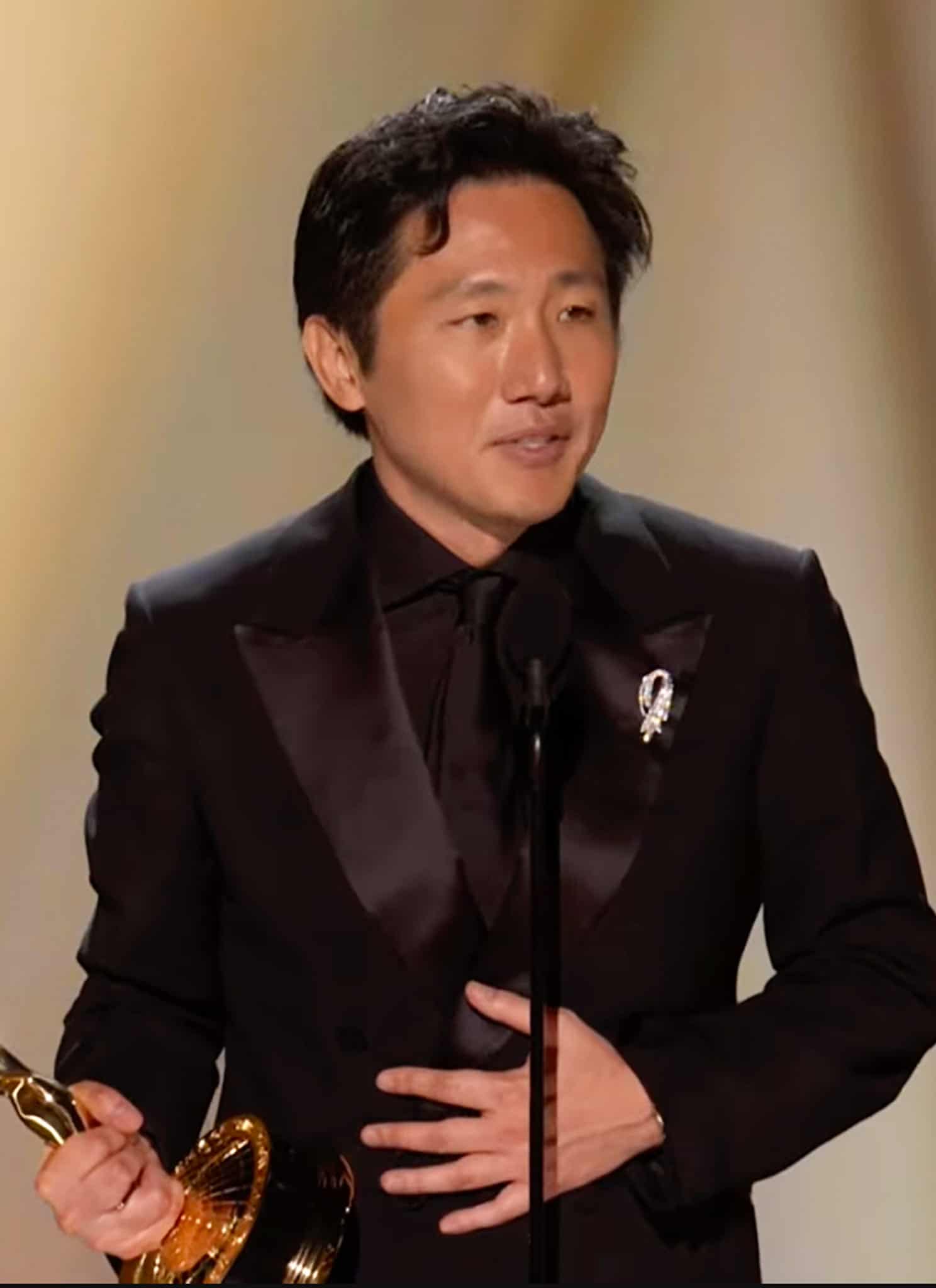 A man in a black suit stands on stage holding an Emmy award, speaking into a microphone against a softly blurred, gold-toned background.