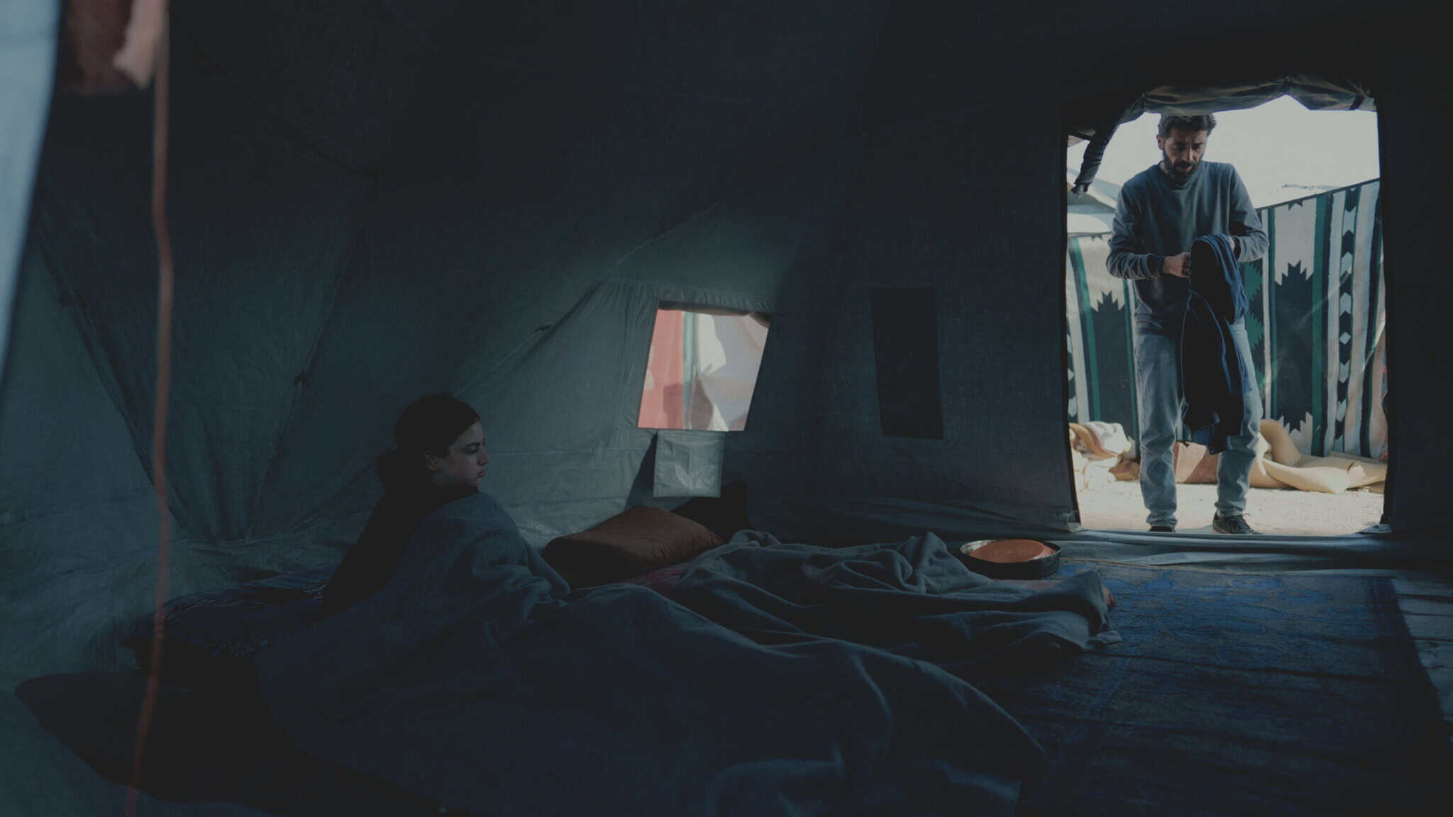 A person lies on a blanket inside a dimly lit tent while another person stands in the doorway, holding a rope or cord. Light enters through the open flap, illuminating part of the scene.