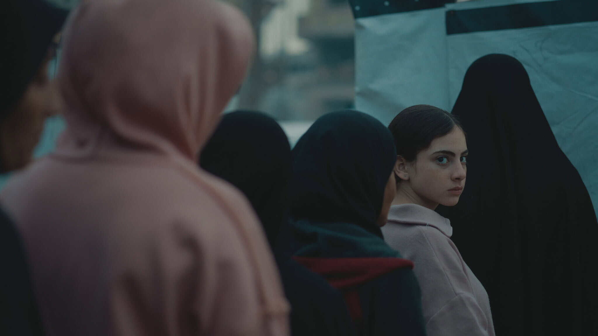 A young woman in a pale hoodie looks over her shoulder amid a group of women wearing dark and light hijabs, standing outdoors near a white tent-like structure.