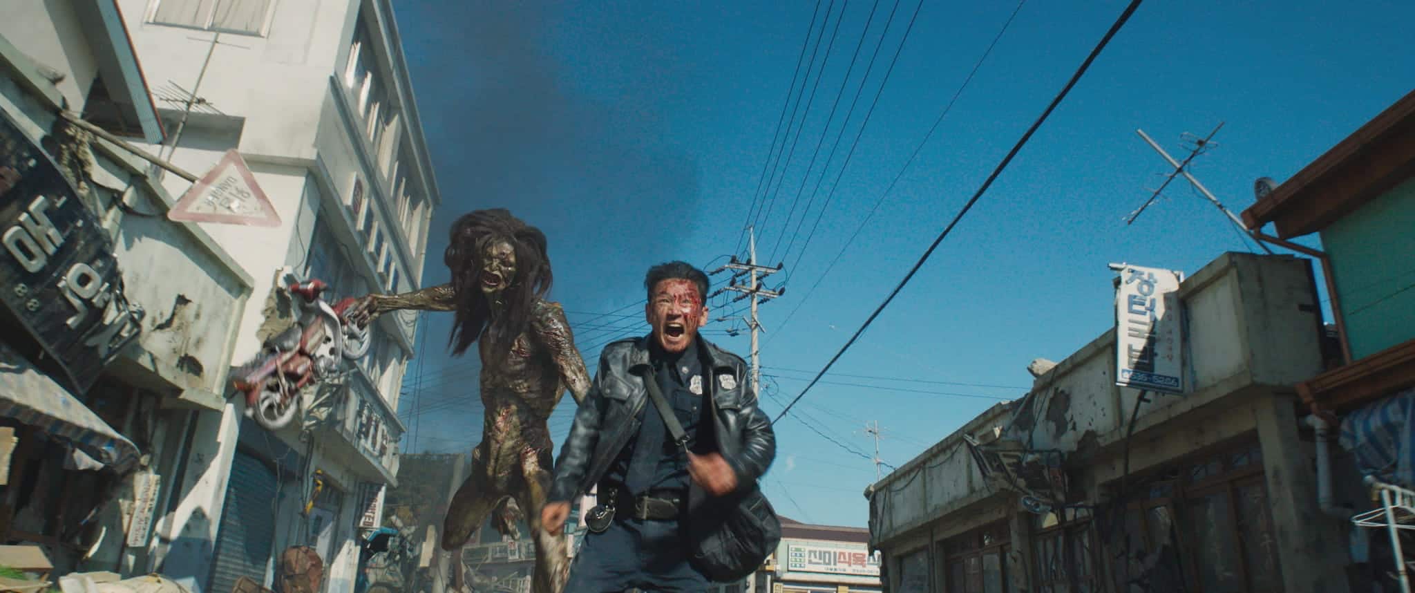 A man in a leather jacket runs screaming down a damaged city street as a monstrous creature with long hair and claws chases him under a clear blue sky.