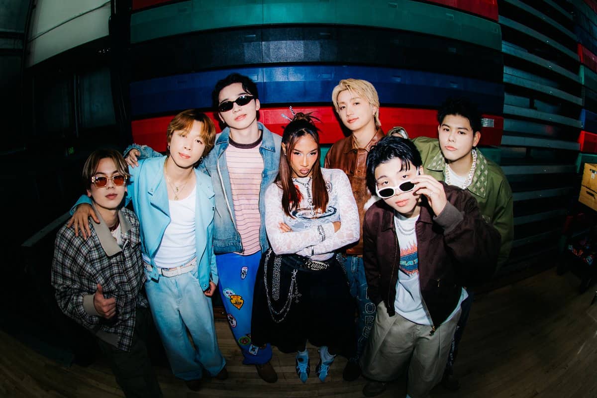 Seven people pose together indoors, dressed in colorful, trendy outfits. Some wear sunglasses and jewelry, and they stand closely, some with arms around each other, against a background of multicolored horizontal panels.