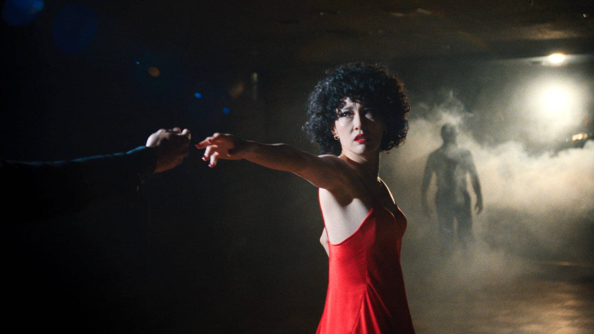 A woman in a red dress reaches out to hold someones hand. She stands in a dimly lit, smoky room, looking intently toward the camera. A shadowy figure is visible in the background.