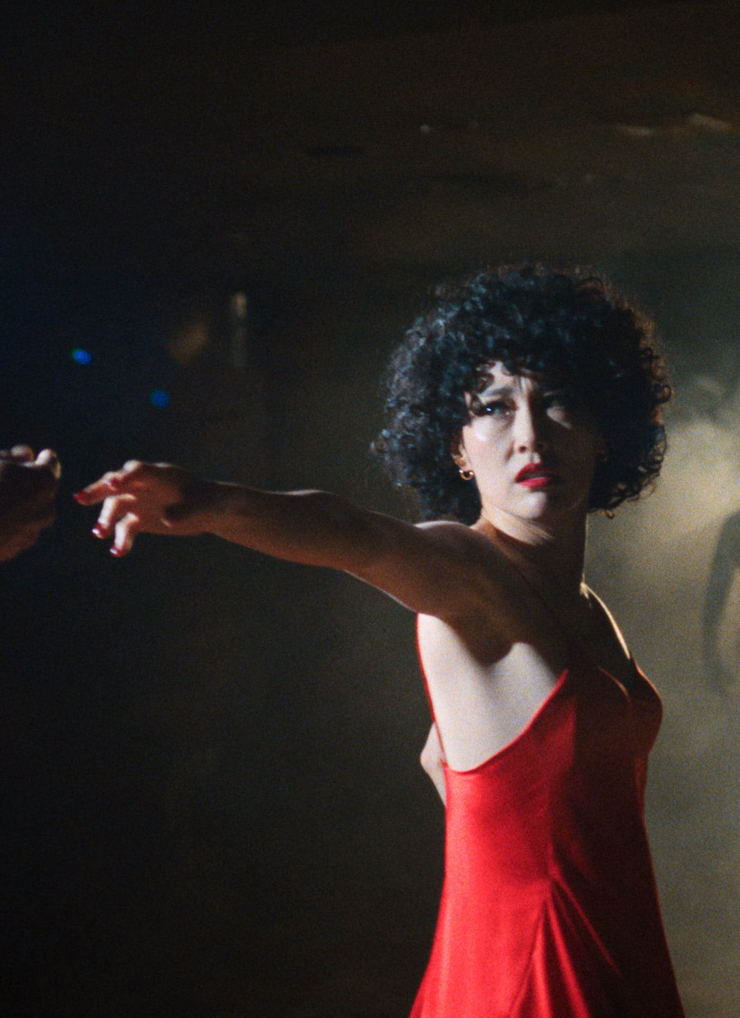 A woman in a red dress reaches out to hold someones hand. She stands in a dimly lit, smoky room, looking intently toward the camera. A shadowy figure is visible in the background.