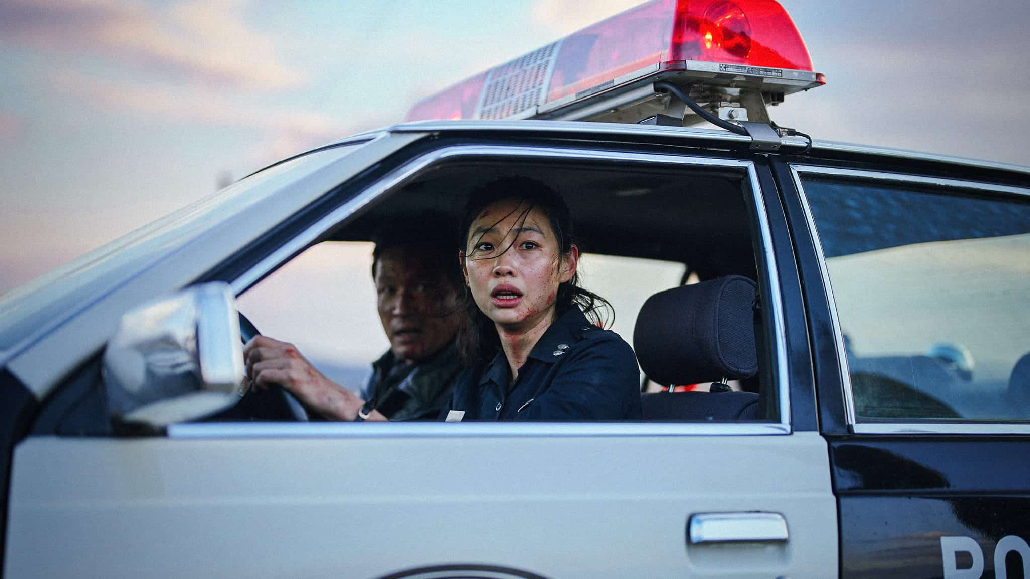 Two police officers sit in a patrol car with the red lights flashing. The woman in the passenger seat looks surprised or alarmed, while the man drives. The setting appears tense, possibly during an emergency or urgent situation.