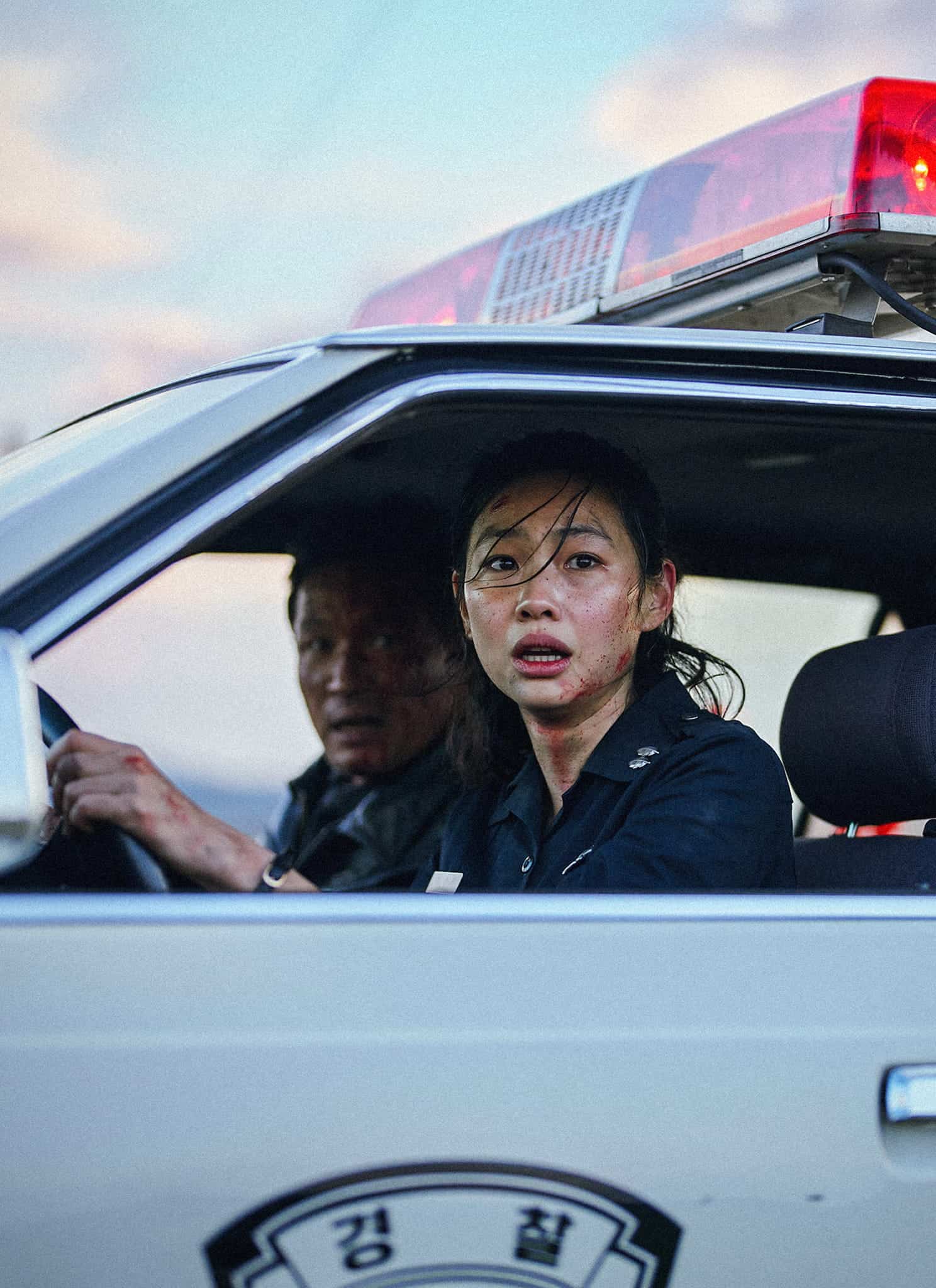 Two police officers sit in a patrol car with the red lights flashing. The woman in the passenger seat looks surprised or alarmed, while the man drives. The setting appears tense, possibly during an emergency or urgent situation.