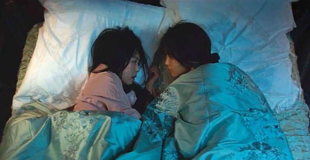 Two women lying in bed facing each other, tucked under a blue patterned duvet.
