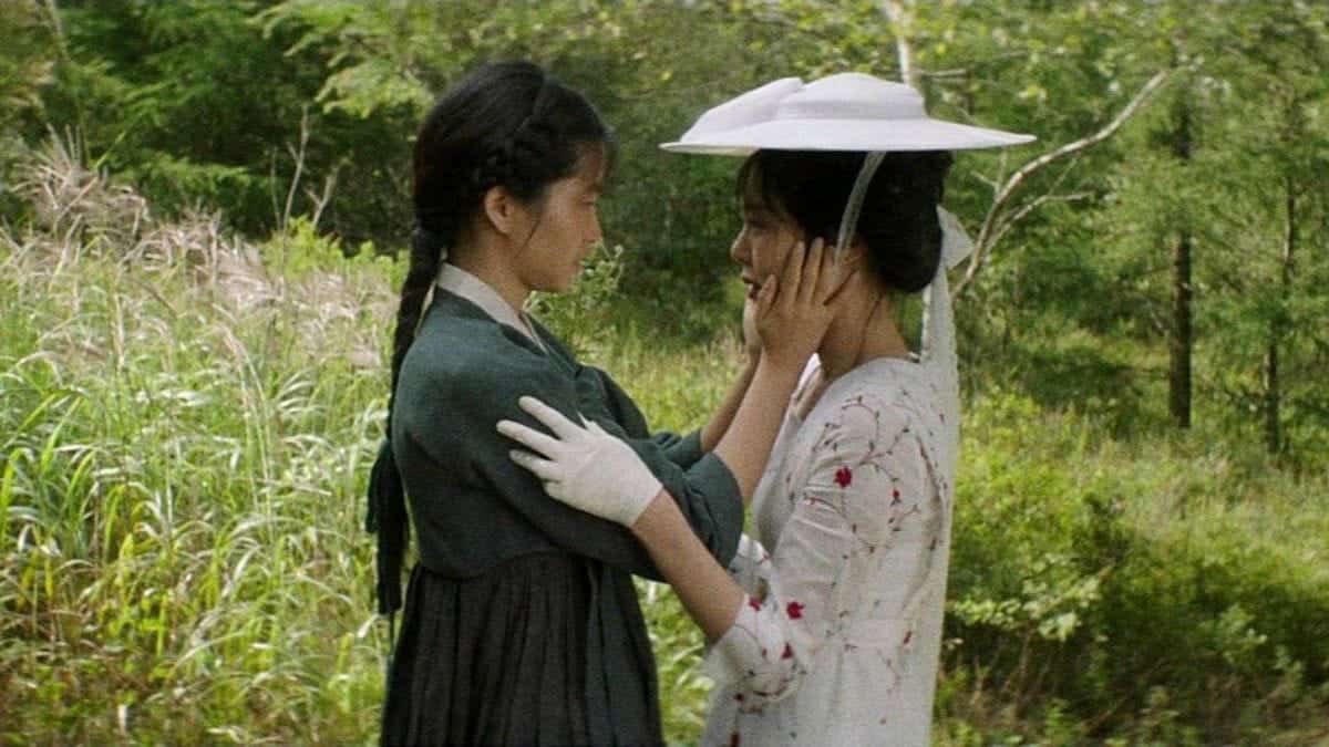 Two women in period clothing stand facing each other in a forest, one wearing white gloves gently touching the others face.