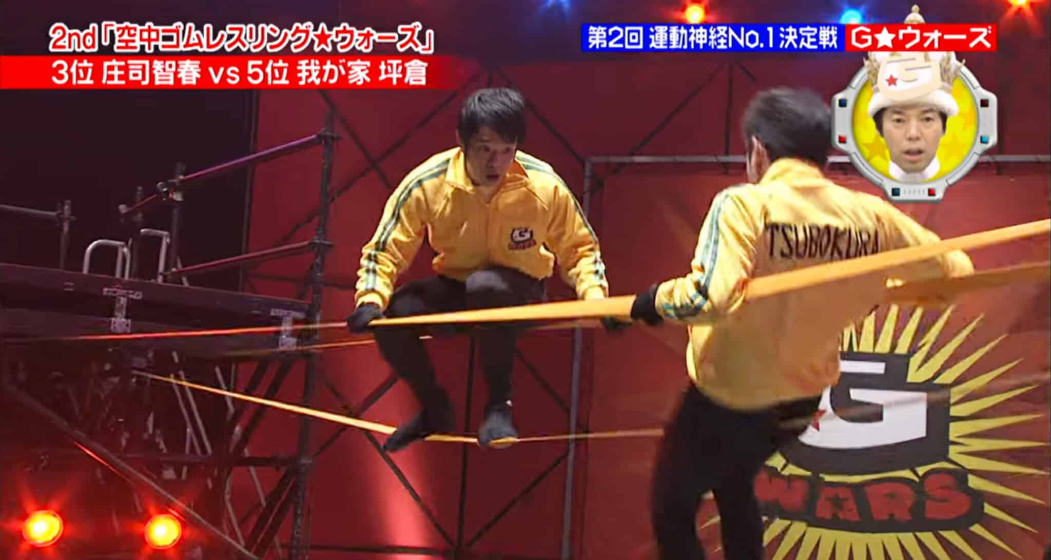 Two contestants in yellow jackets balance and compete on parallel slacklines in a brightly lit indoor arena, with a hosts face shown in the corner and Japanese text displayed across the screen.