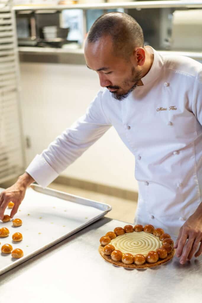 A chef in a white uniform arranges caramel-coated pastries on a tart in a professional kitchen, focusing on placing the pastries in a circular pattern around the dessert.