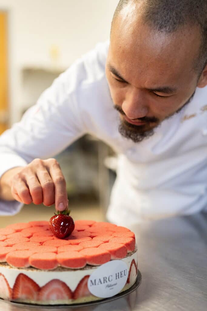 A pastry chef in a white coat carefully places a strawberry on top of a strawberry cake decorated with sliced strawberries and a MARC HEU label.