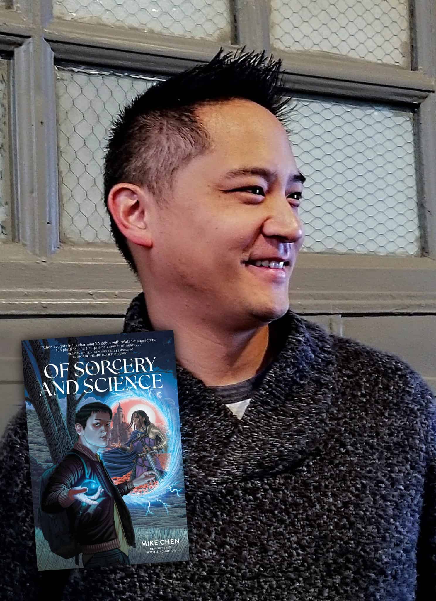 A smiling man in a gray sweater stands in front of a window. Next to him is the cover of the book Of Sorcery and Science by Mike Chen, featuring a young man holding a glowing device and a woman with magical energy.