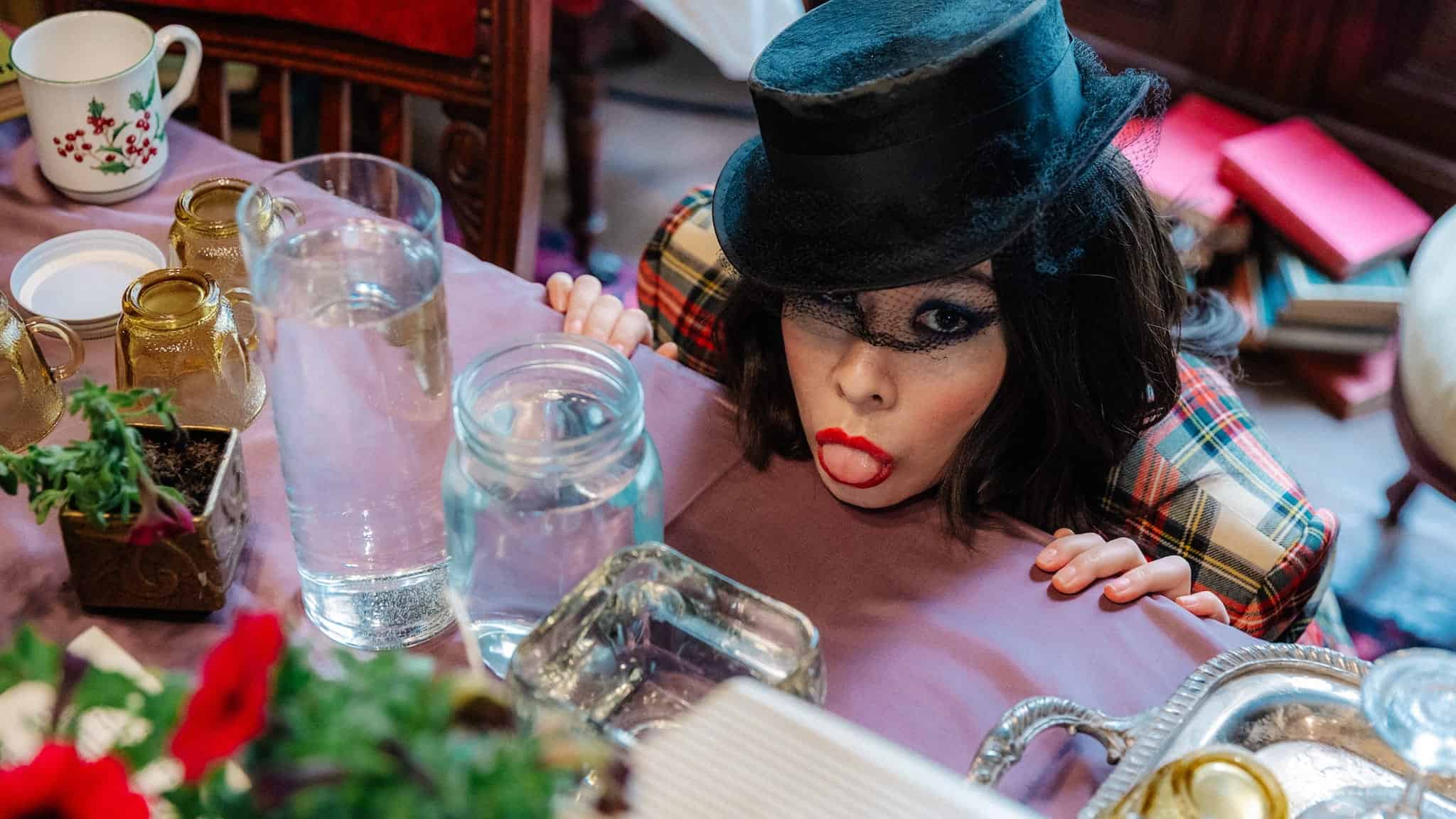 A person in a plaid outfit and black top hat playfully sticks out their tongue while peeking over a table set with glassware, cups, and flowers, channeling serious Mitski energy.