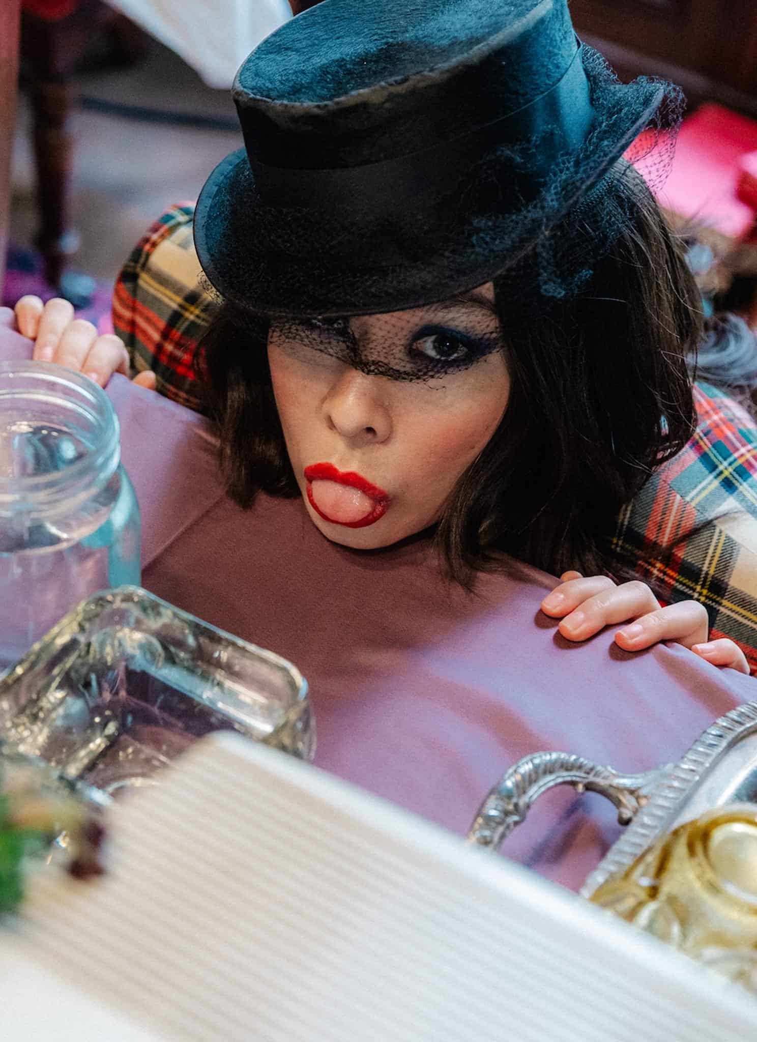 A person in a plaid outfit and black top hat playfully sticks out their tongue while peeking over a table set with glassware, cups, and flowers, channeling serious Mitski energy.