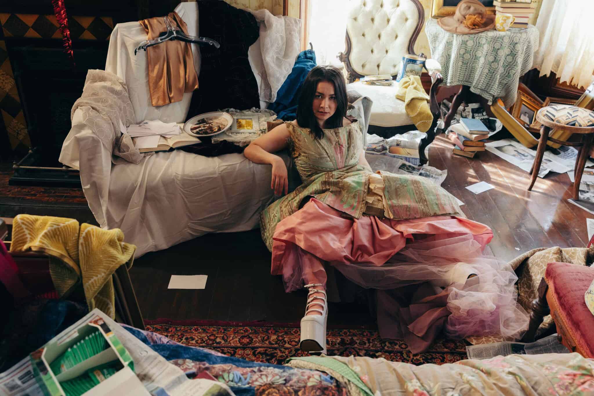A woman in a colorful dress sits on a sofa in a cluttered room with scattered clothes, papers, and food containers, looking at the camera—her expression reminiscent of a Mitski album cover, capturing both longing and quiet chaos.