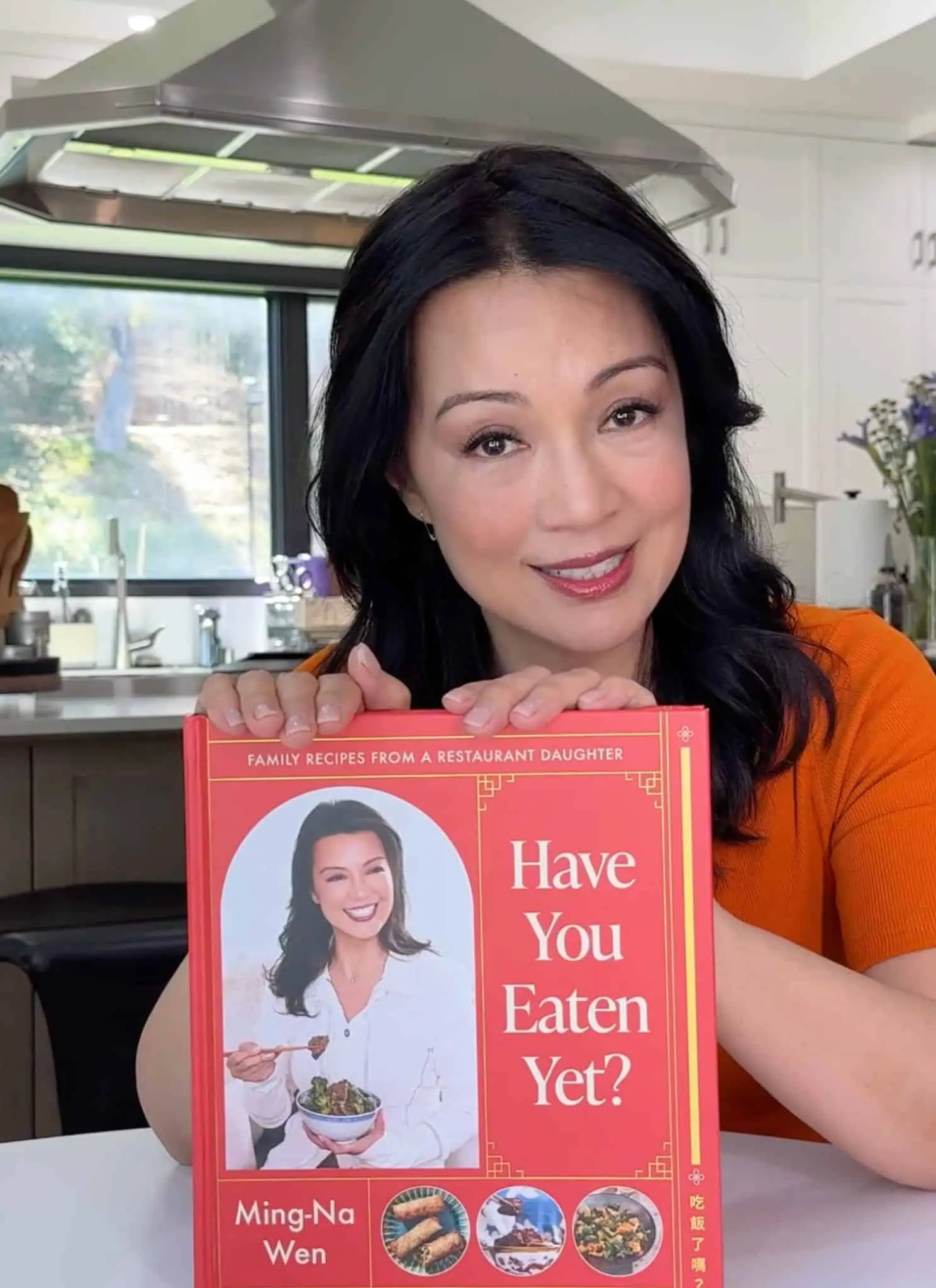 Ming-Na Wen smiles while holding her cookbook, Have You Eaten Yet?, in a bright kitchen.