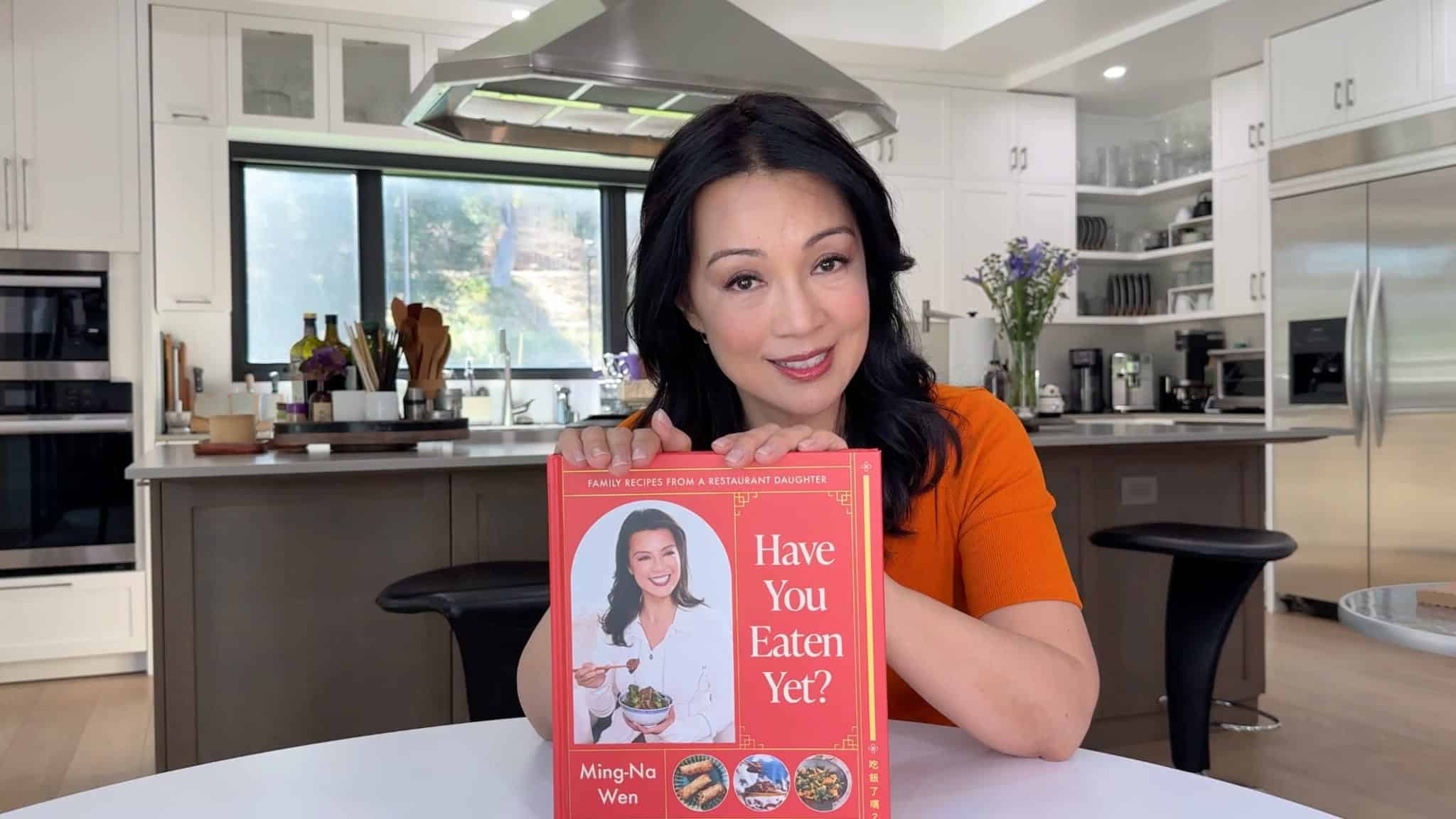 Ming-Na Wen smiles while holding her cookbook, Have You Eaten Yet?, in a bright kitchen.