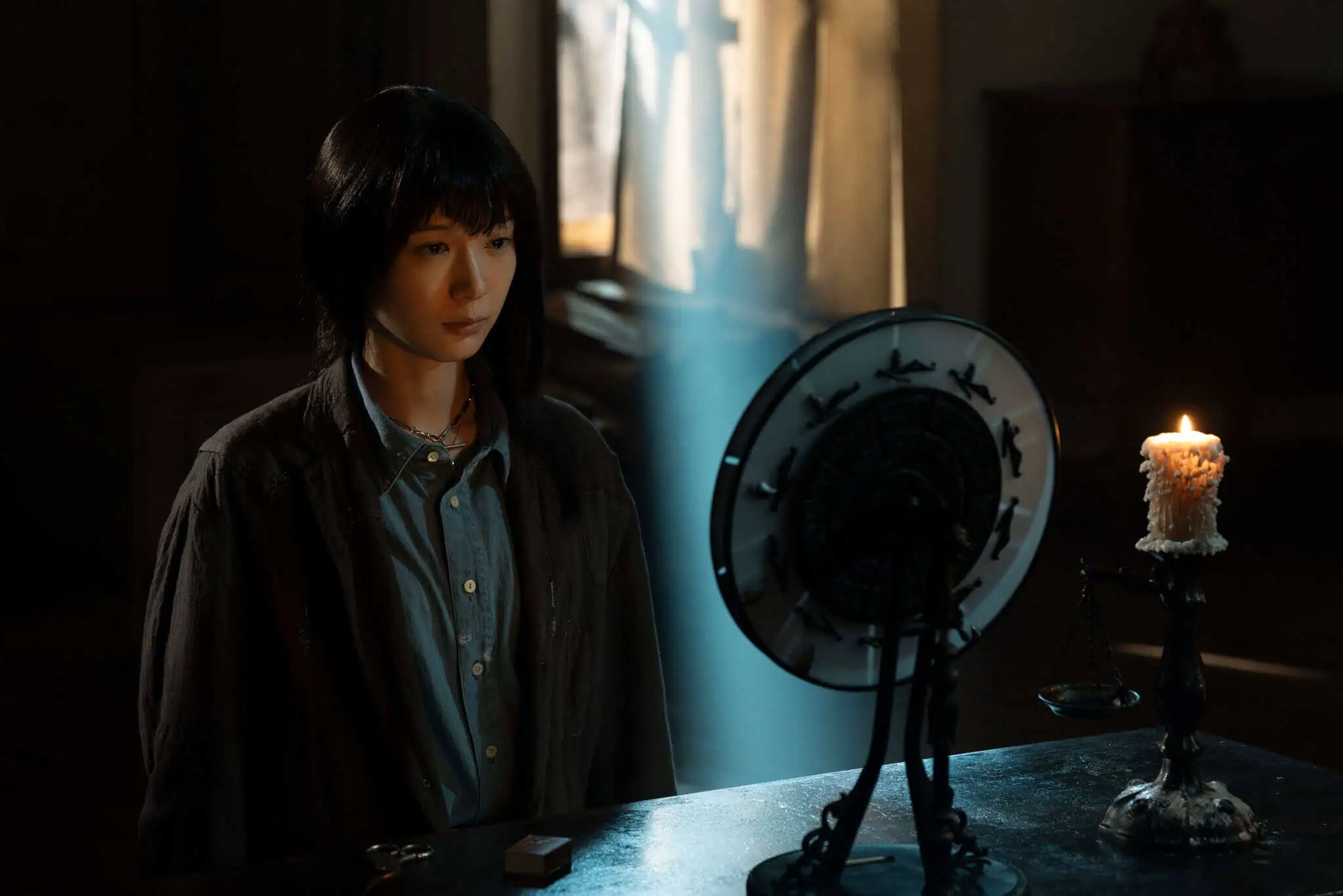 A woman with dark hair sits at a table facing a round mirror, with a lit candle and a scale beside her. Sunlight streams through a window, casting a blue beam across the dimly lit room.