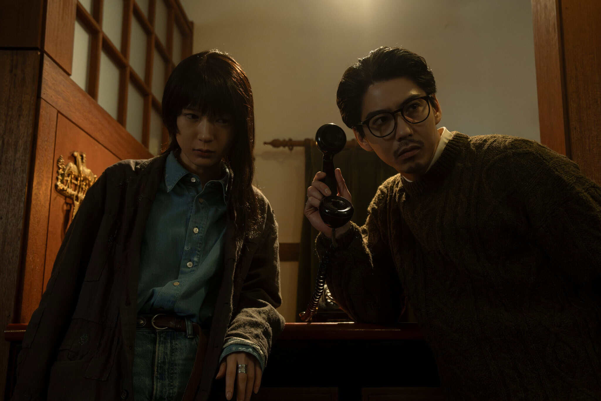 A man holds an old-fashioned phone to his ear while looking serious; beside him, a woman in layered clothing leans against a counter, also appearing tense. They are in a dimly lit, wood-paneled room.