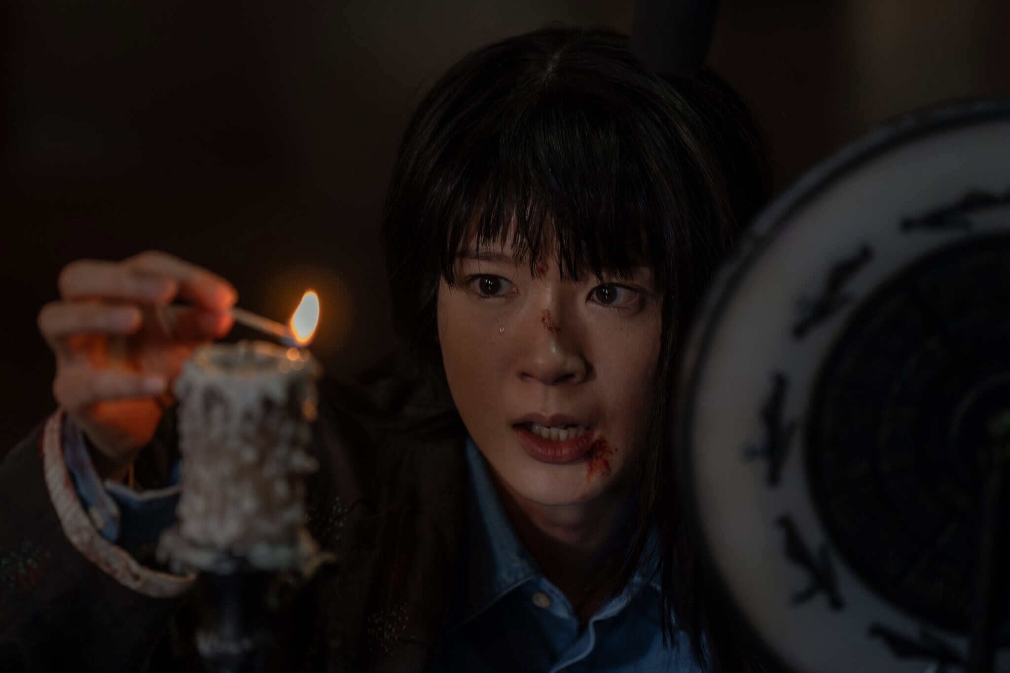 A woman with a bruised face lights a candle with a match. She looks intensely focused and anxious, with a round, patterned object partially visible beside her.