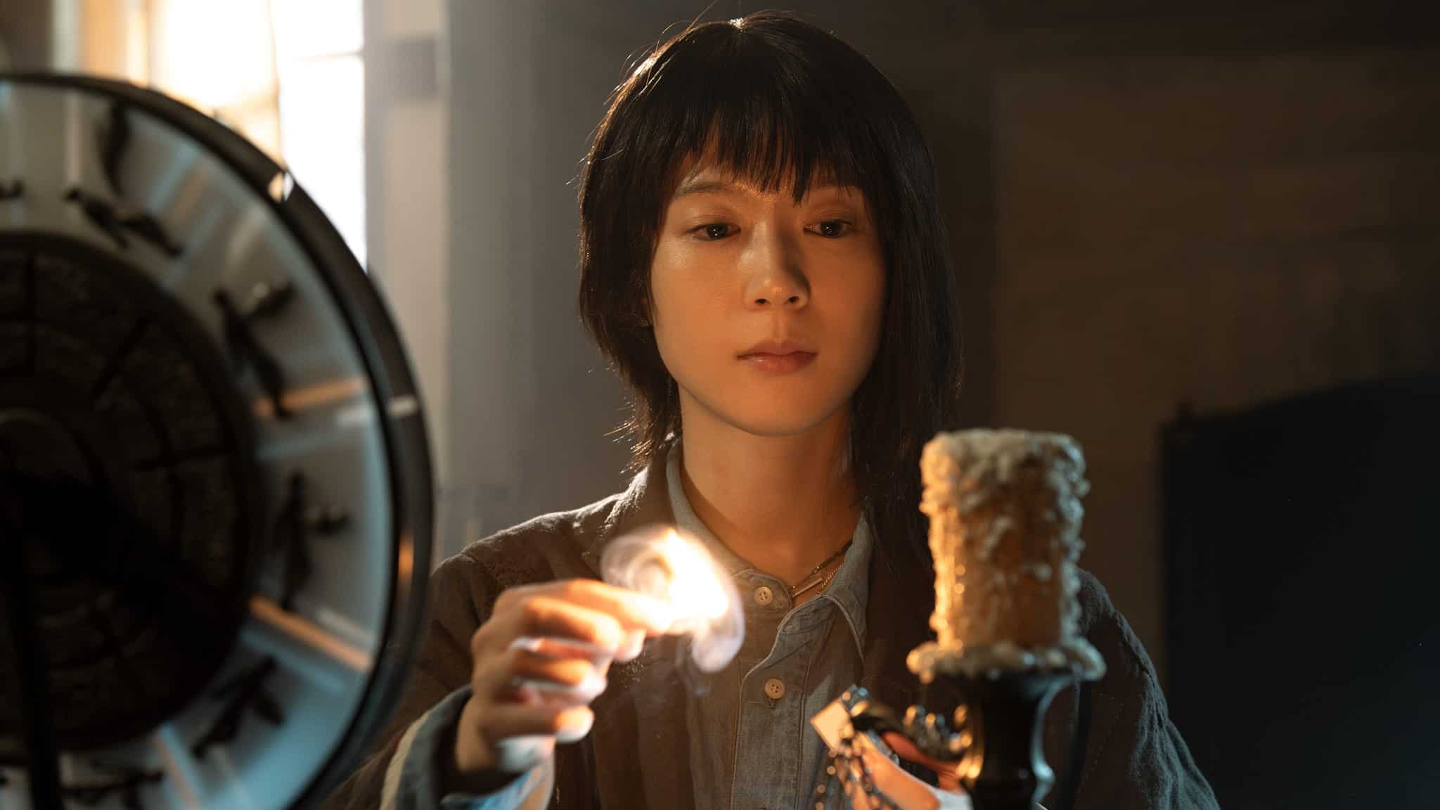 A person with short dark hair lights a worn, textured candle using a match, with sunlight streaming through a window and a large circular object nearby.