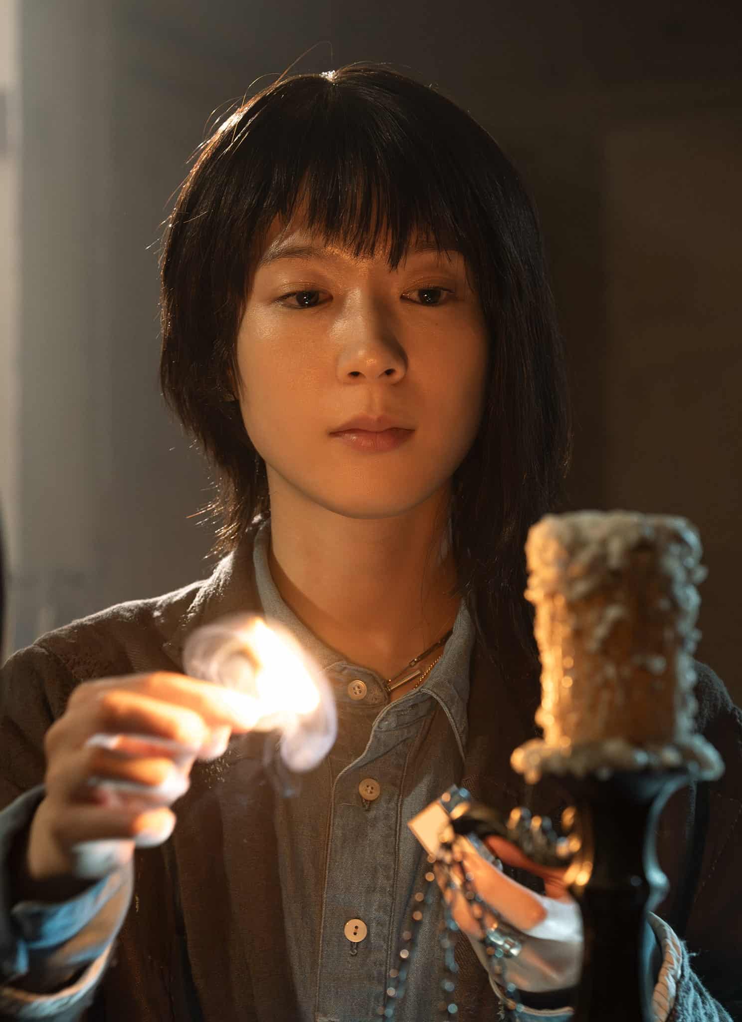 A person with short dark hair lights a worn, textured candle using a match, with sunlight streaming through a window and a large circular object nearby.