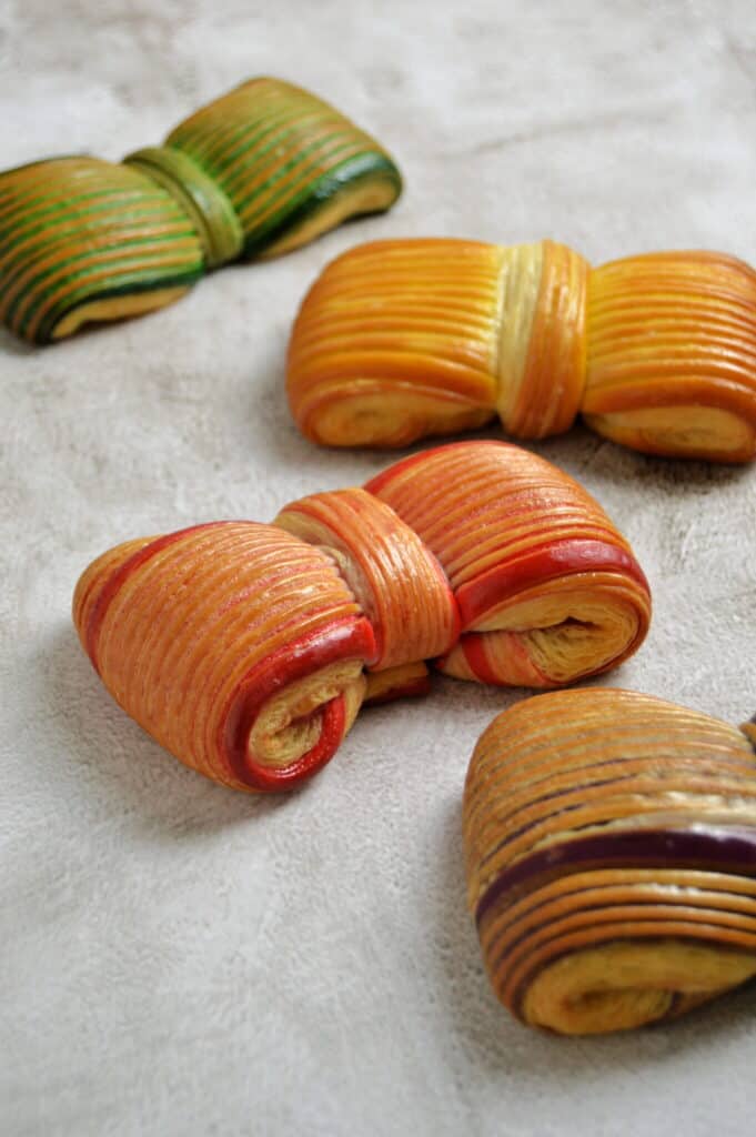 Four colorful, folded pastries with visible layers are arranged on a light textured surface. Each pastry has distinct hues, including green, yellow, orange, and red tones.