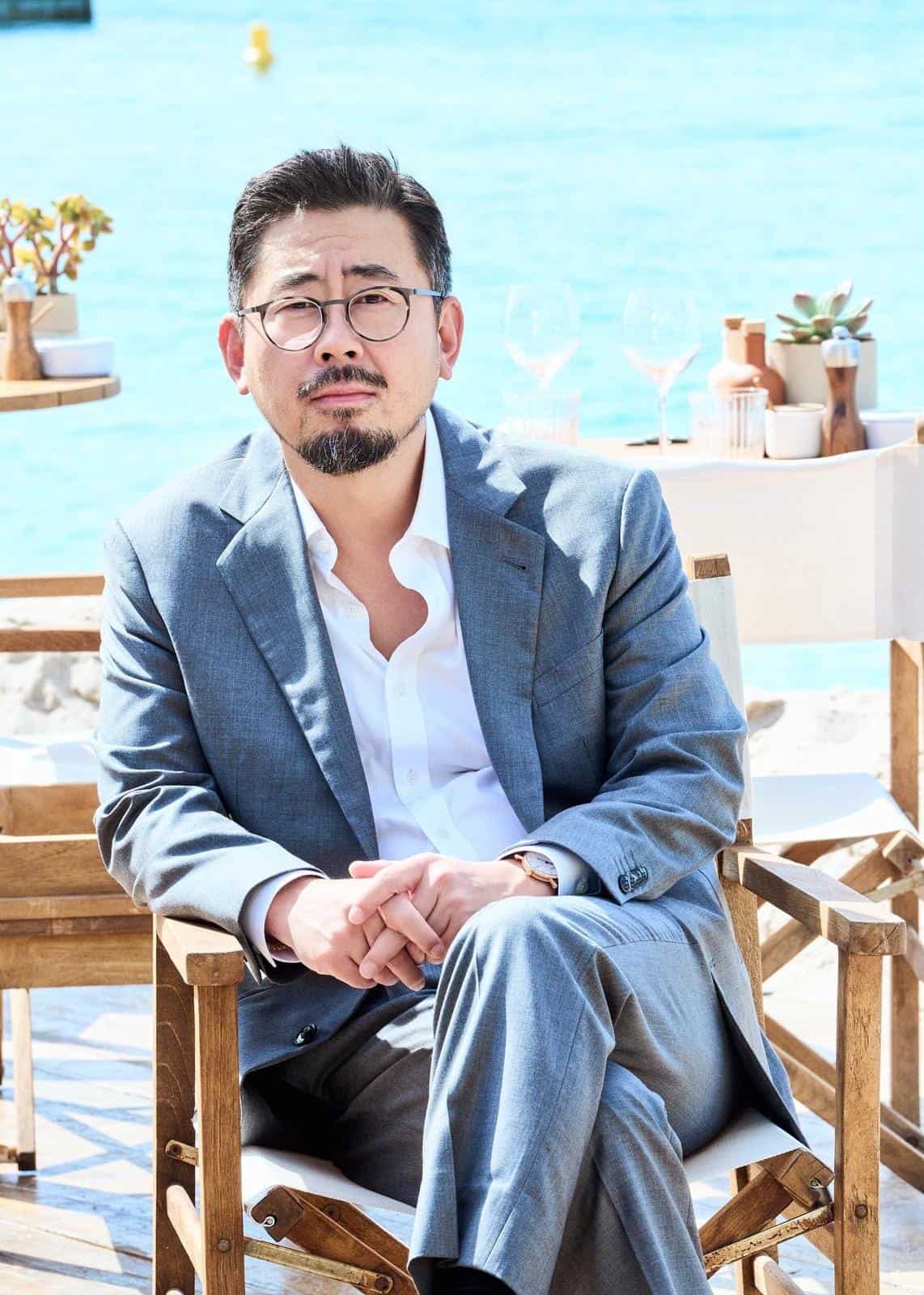 A man in a gray suit and white shirt sits on a wooden chair outdoors by the water, with potted plants and tables in the background, evoking the cinematic atmosphere often found in Na Hong-jin’s films.
