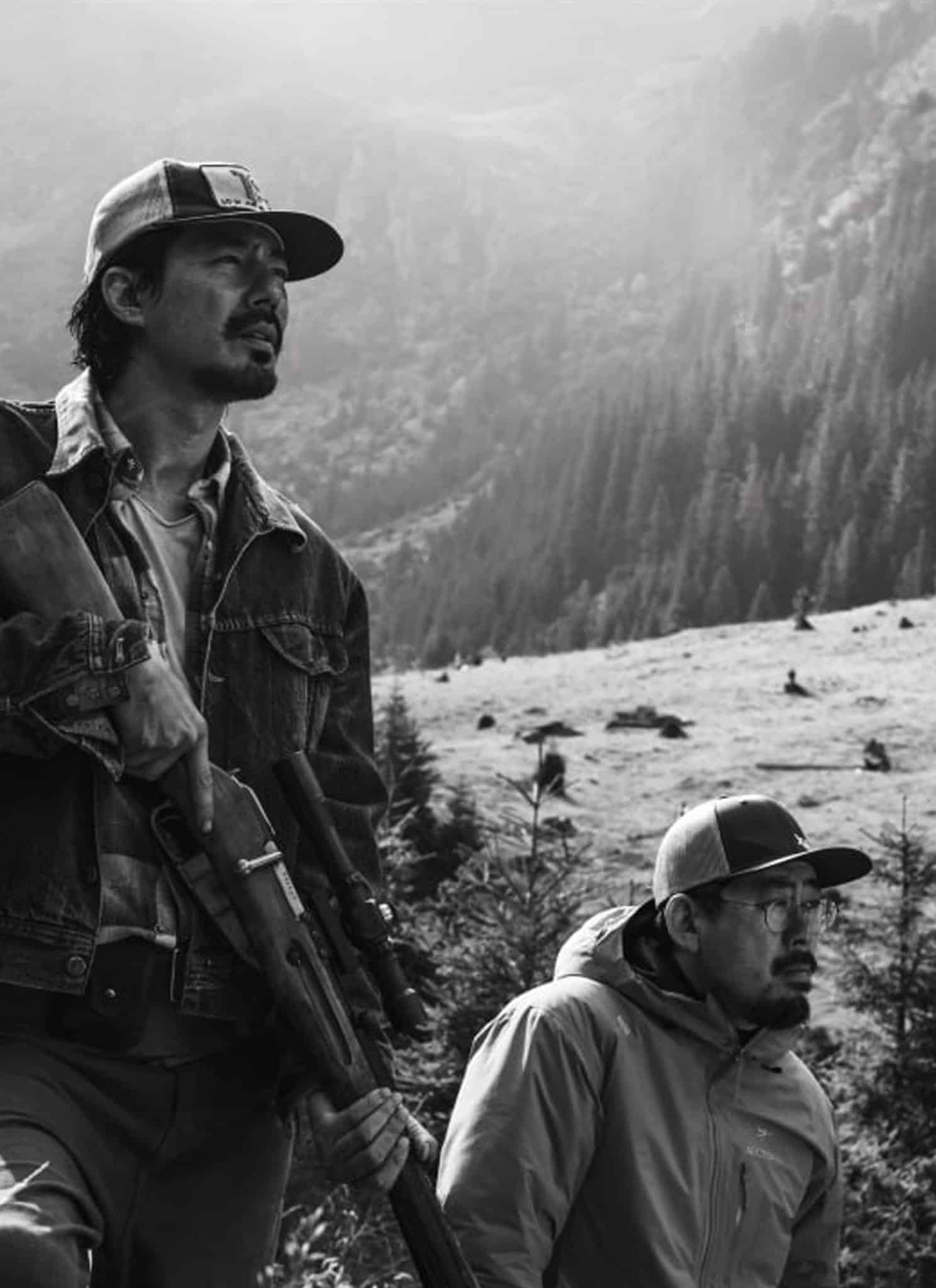 Two people in outdoor clothing and hats walk through a mountainous, forested area. One holds a rifle. The scene is in black and white, evoking a remote and rugged landscape reminiscent of the atmospheric tension found in Na Hong-jin’s films.