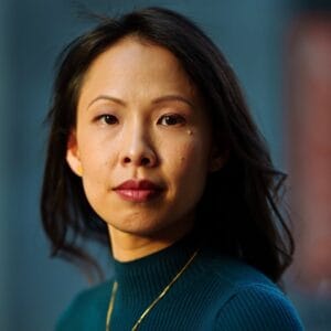 A woman with medium-length dark hair wears a teal sweater and a gold necklace, looking directly at the camera with a neutral expression. The background is softly blurred in blue tones.