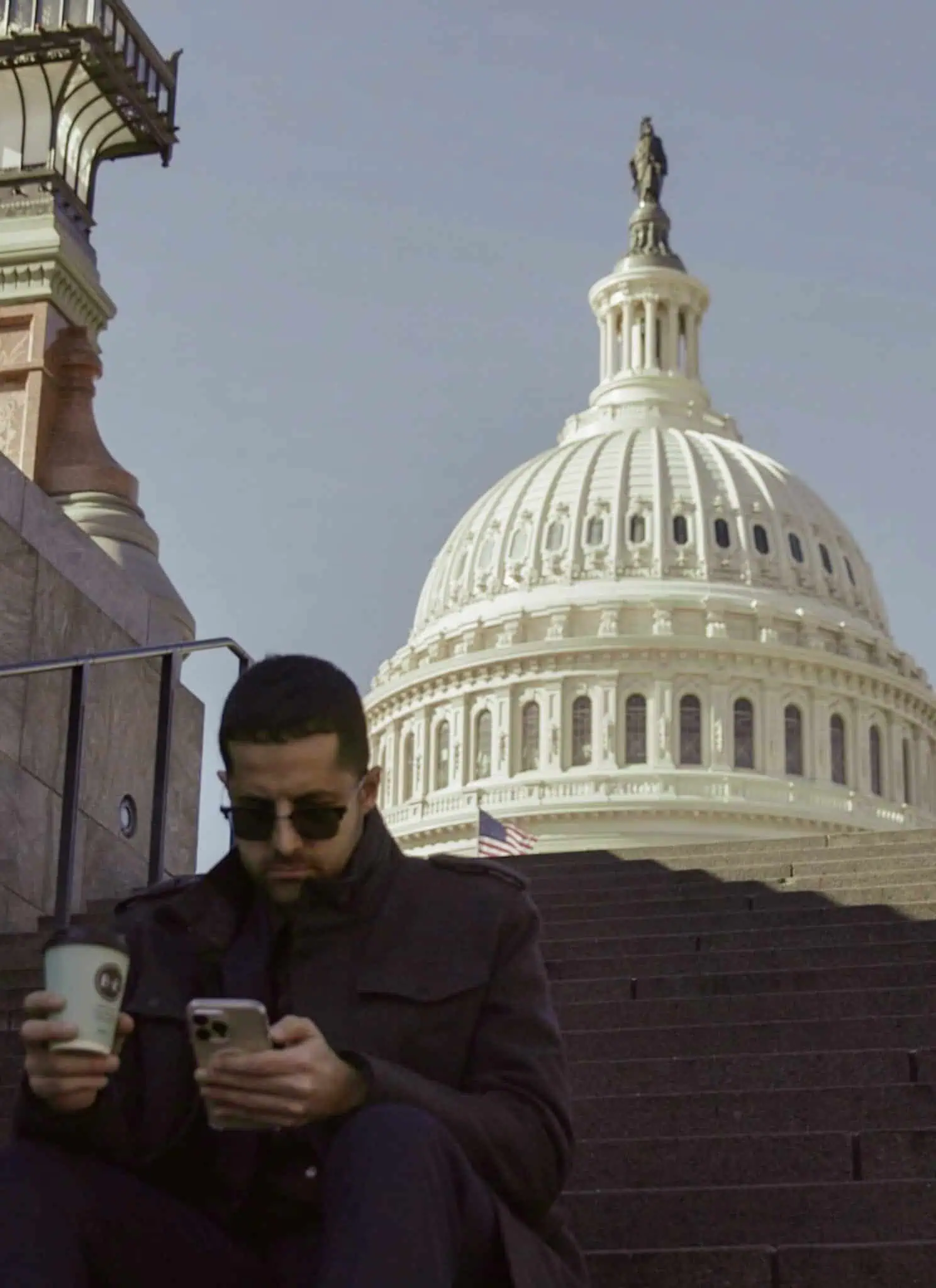 A man sits on steps holding a coffee cup and phone, looking at his phone. The U.S. Capitol dome rises in the background under a clear sky.