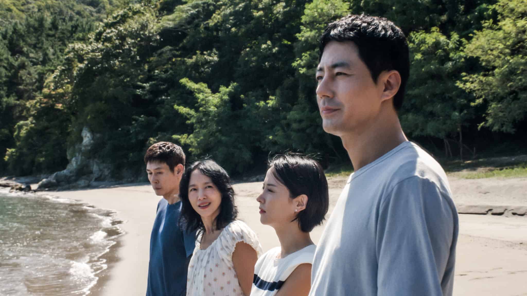 Four people stand on a sandy beach near the water, with trees and greenery in the background. They are looking in different directions, some gazing at the sea and others at each other. The scene appears calm and thoughtful.