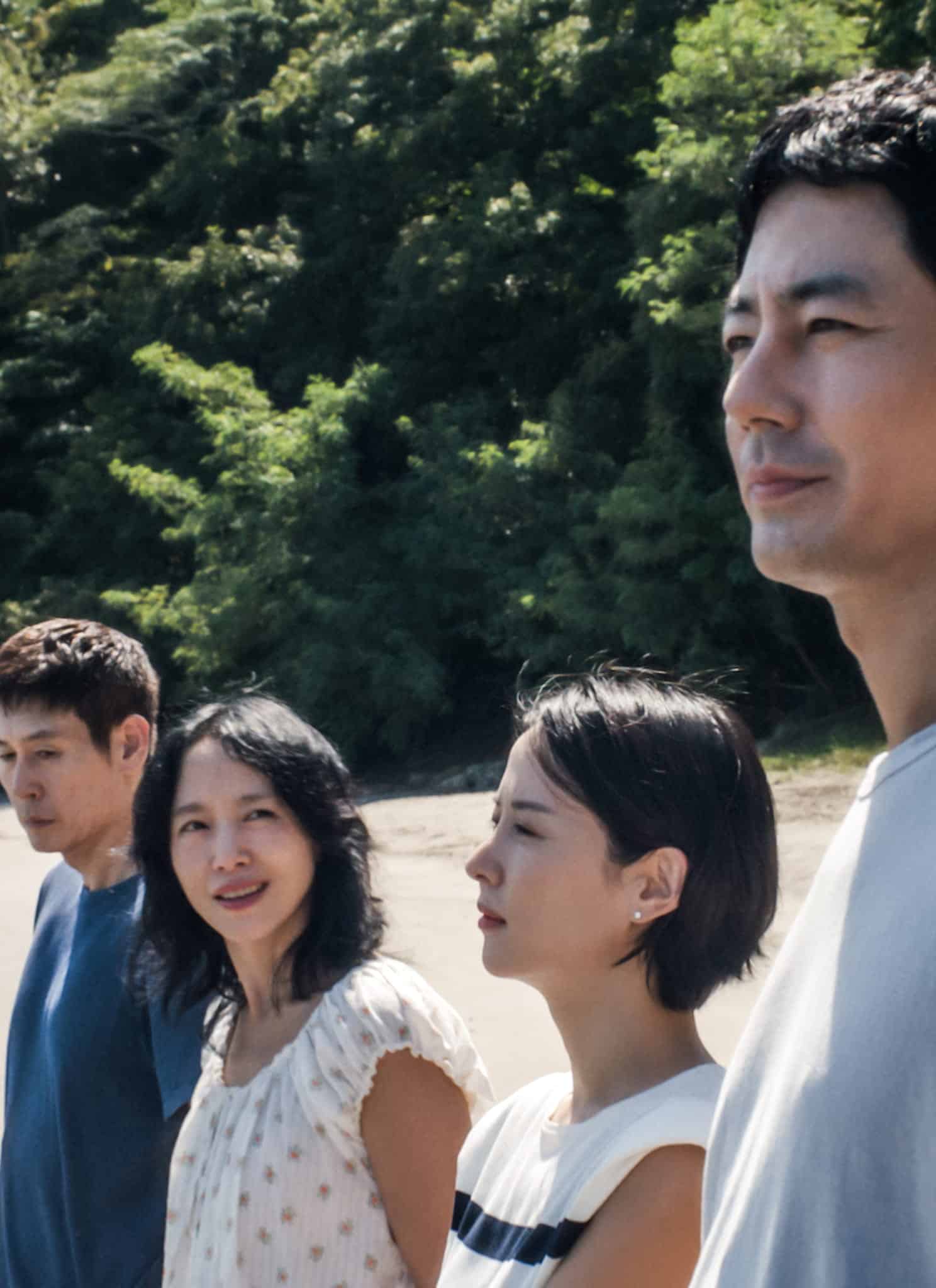 Four people stand on a sandy beach near the water, with trees and greenery in the background. They are looking in different directions, some gazing at the sea and others at each other. The scene appears calm and thoughtful.